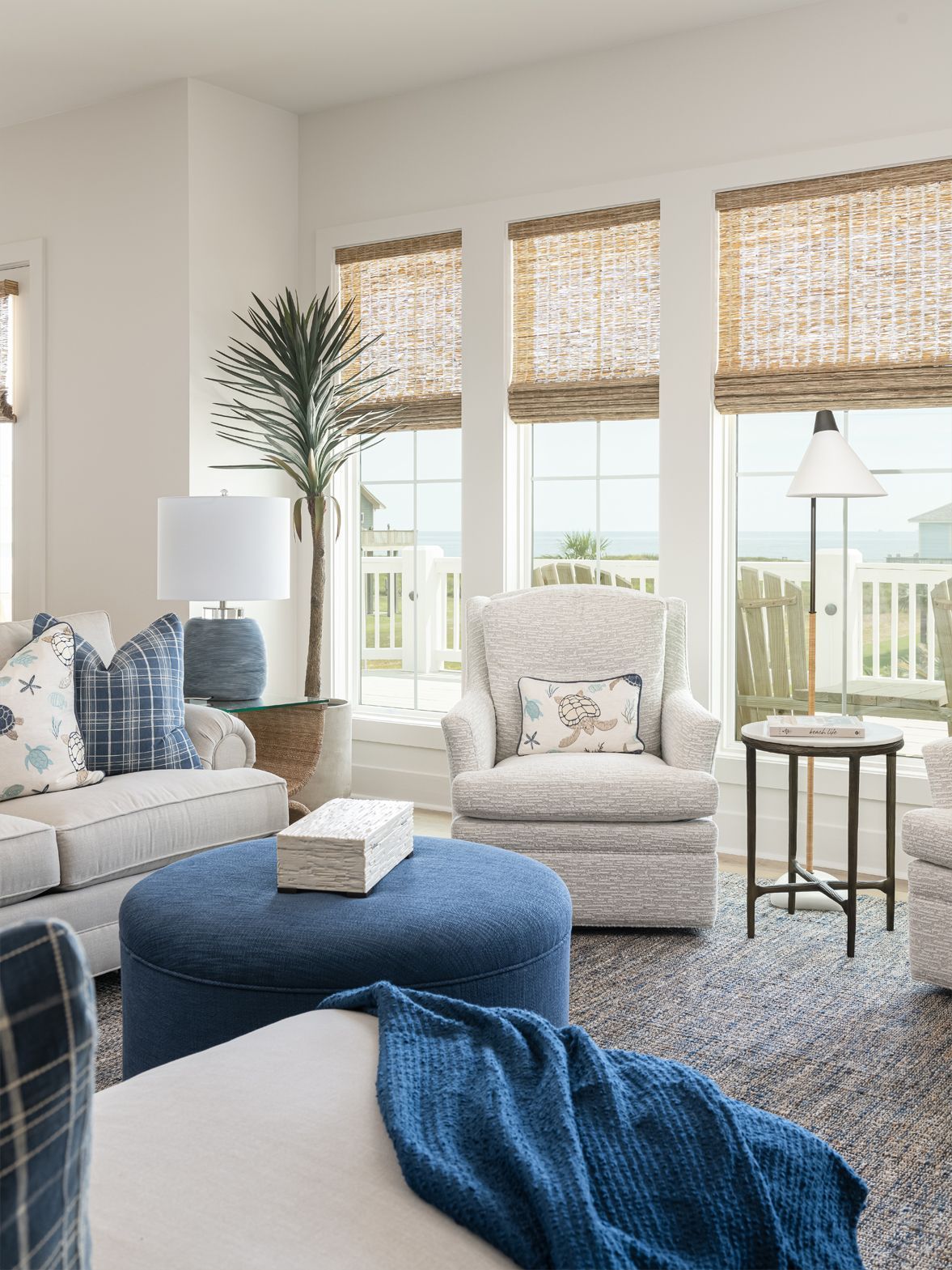 Bright living room with beige furniture, blue accents, woven shades, and large windows overlooking a deck