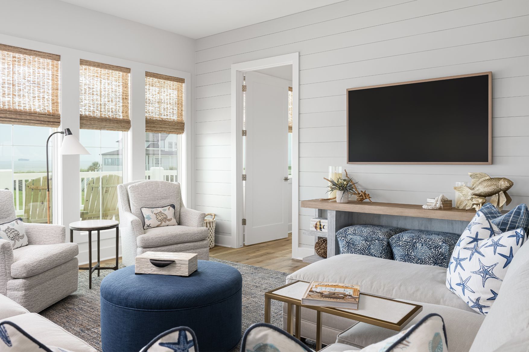 Bright living room with white walls, navy ottoman, armchairs, and a wall-mounted TV above a fireplace.