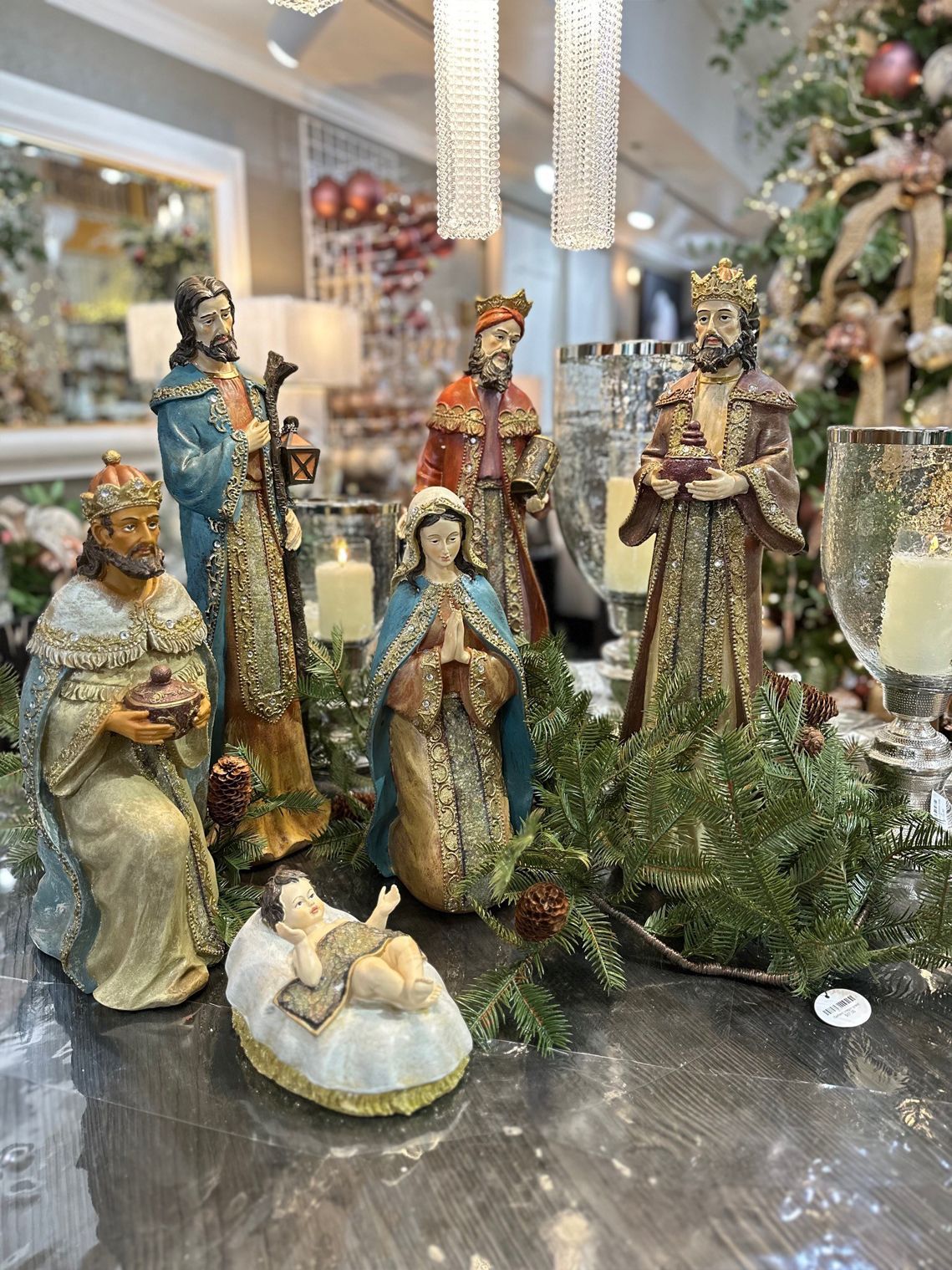 Nativity figurines arranged on a reflective table, surrounded by greenery and holiday decor