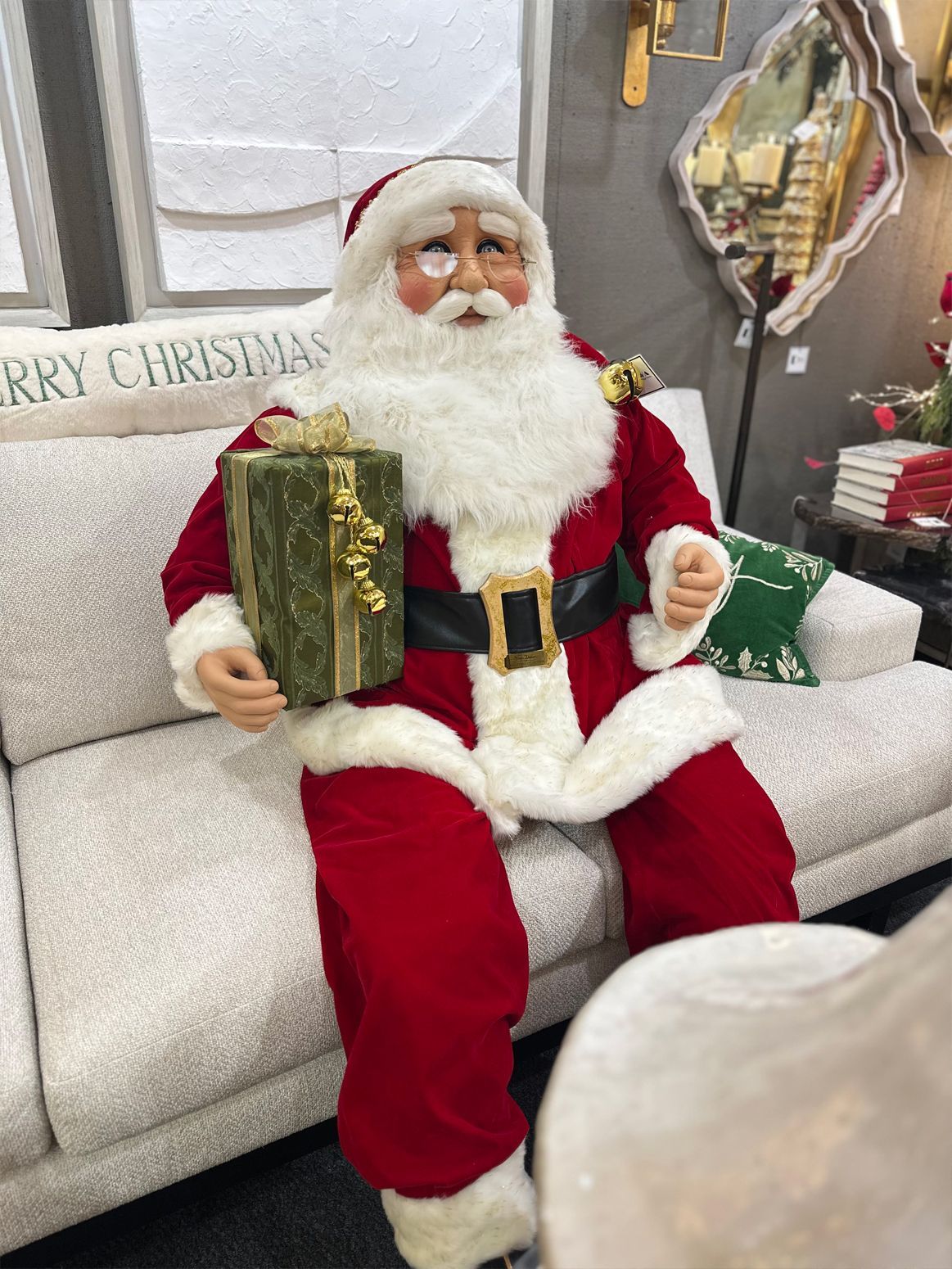 Santa figurine in a red suit holding a gold gift, seated on a white couch with holiday decor in the background