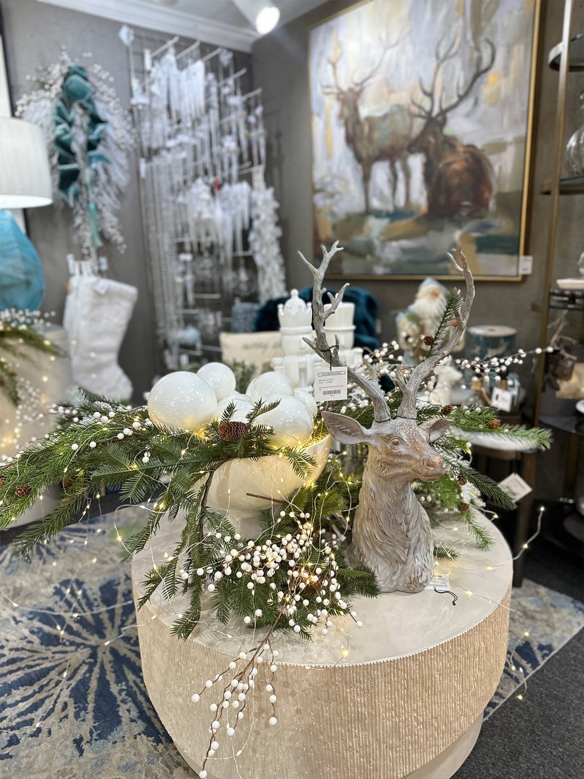 Festive holiday display with white reindeer, pine branches, lights, and silver ornaments on a round table