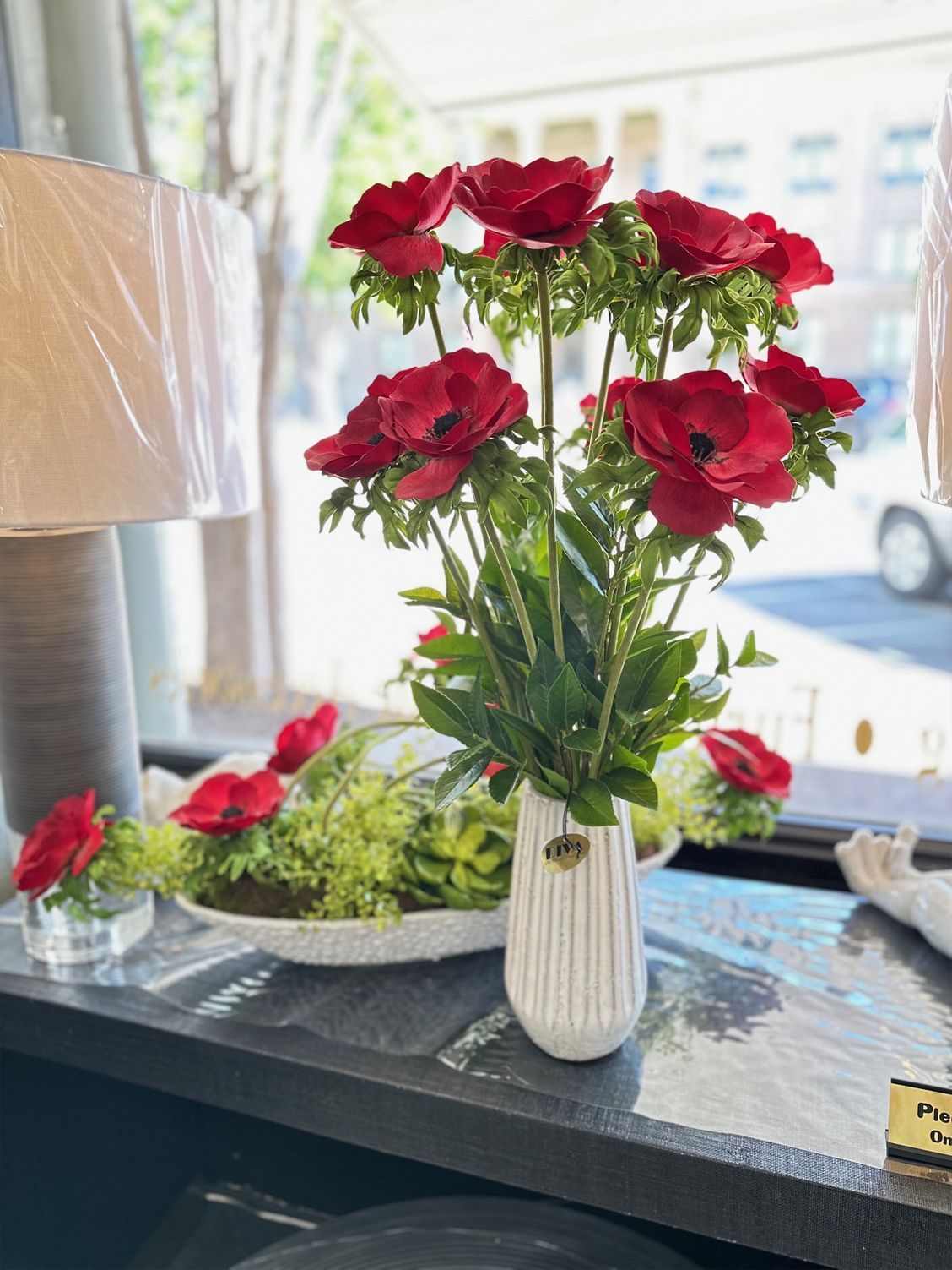 Red flowers in a white vase on a tabletop by a bright window.