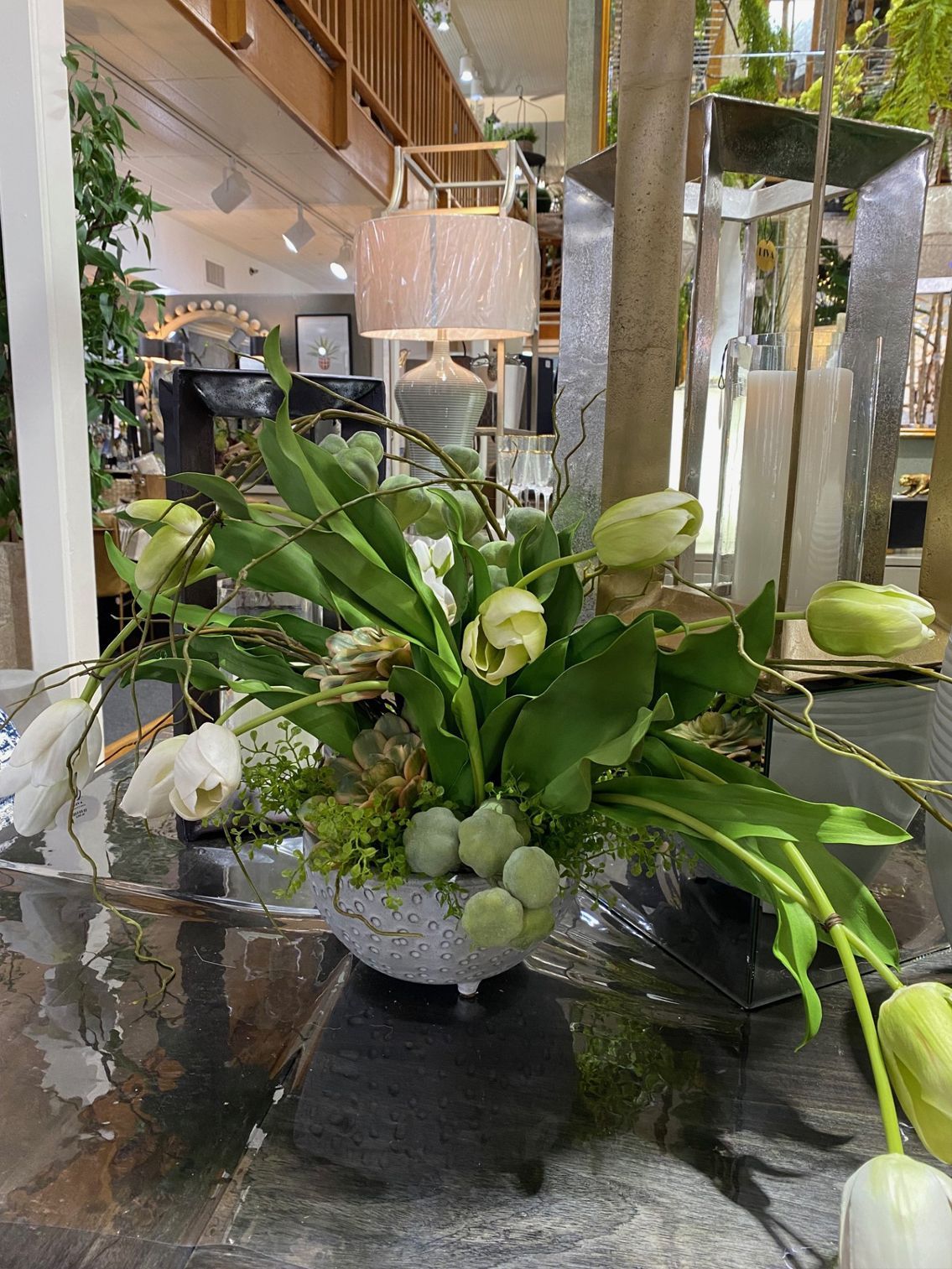 White floral centerpiece with green leaves and stems in a glass vase on a reflective table.