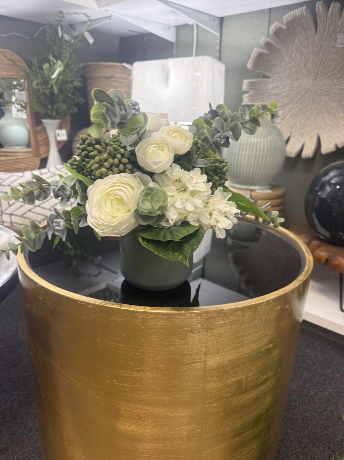 White floral arrangement in a black vase on a gold table, with greenery in a decorative showroom.