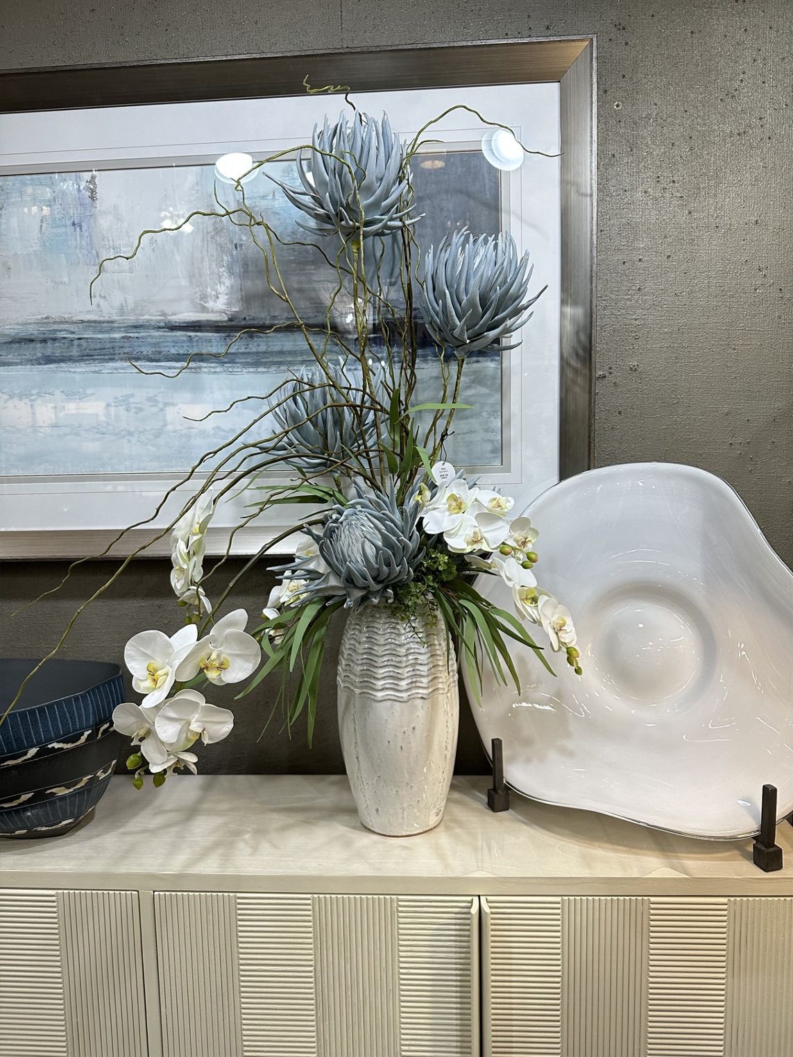 White floral arrangement in a silver vase beside a decorative plate on a mantel.