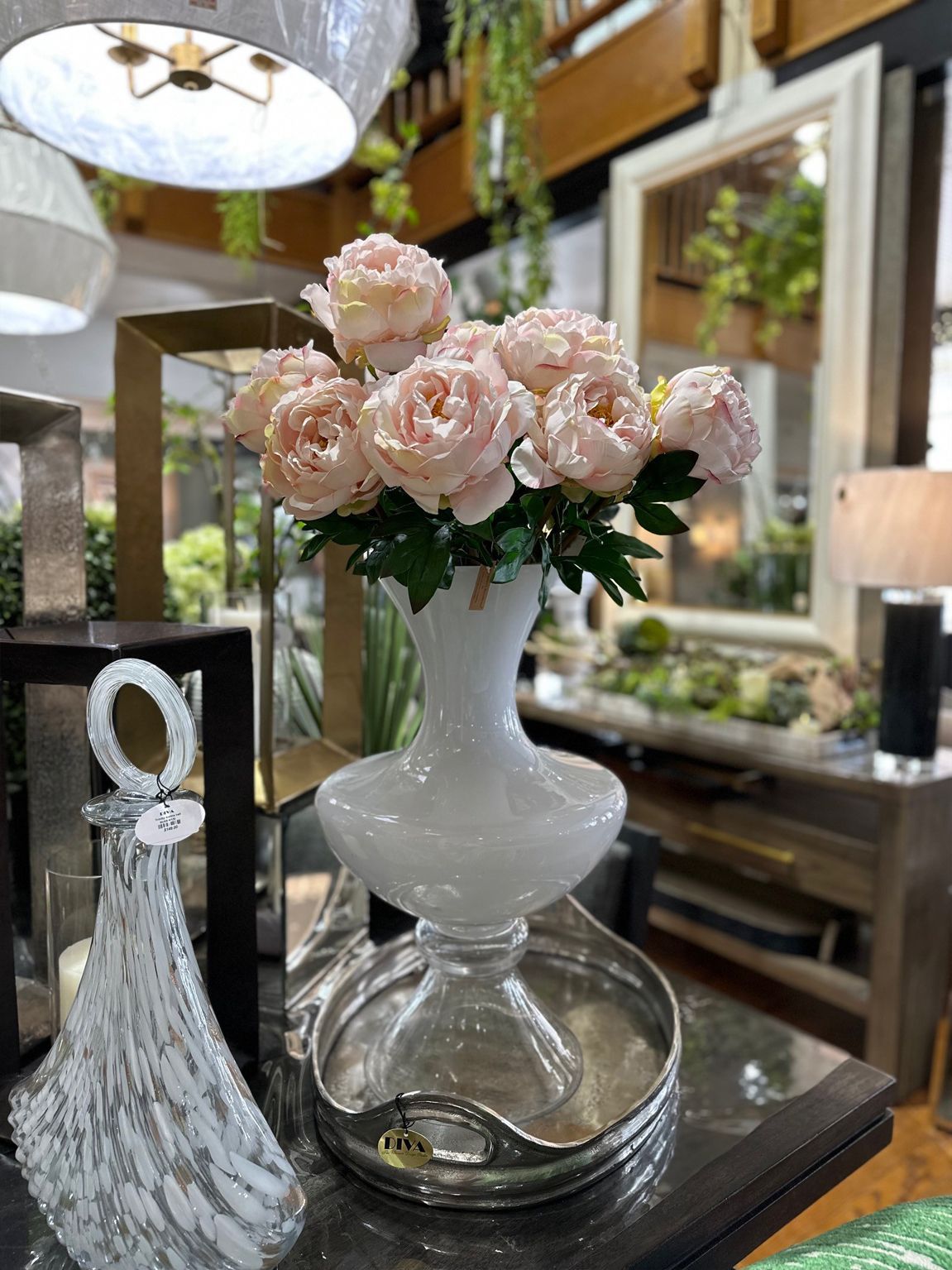 Pink flowers in a white vase on a wooden tray beside a decorative glass bottle in a bright room