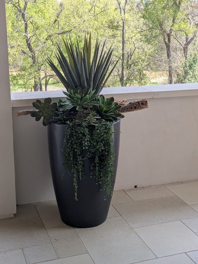Tall black planter with green succulents on a tiled balcony, with trees in the background
