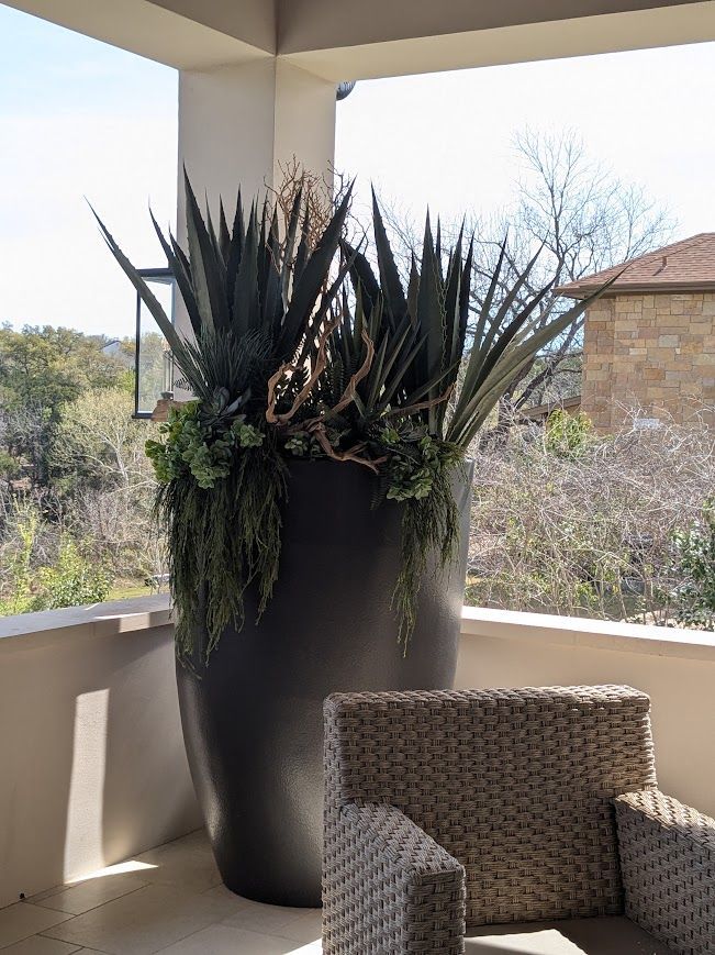 Large black planter with spiky green plants on a balcony beside a woven chair, with desert landscape beyond