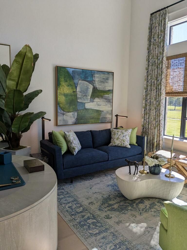 Modern living room with navy sofa, abstract wall art, white coffee table, and tall windows with greenery.