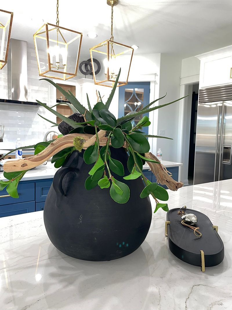 Black vase with leafy plant and decorative branches on a kitchen island, with a small black object beside it