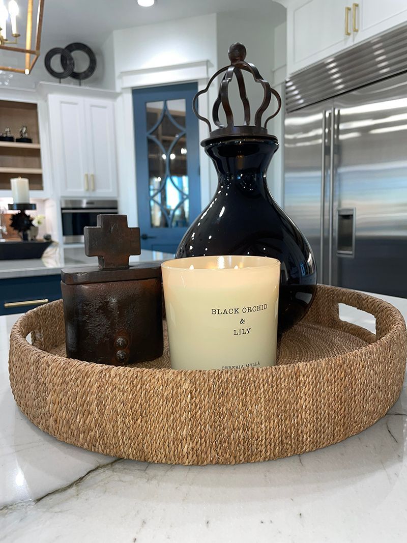 Woven tray with candles and decor on a kitchen island, with a black vase and stainless steel fridge behind.
