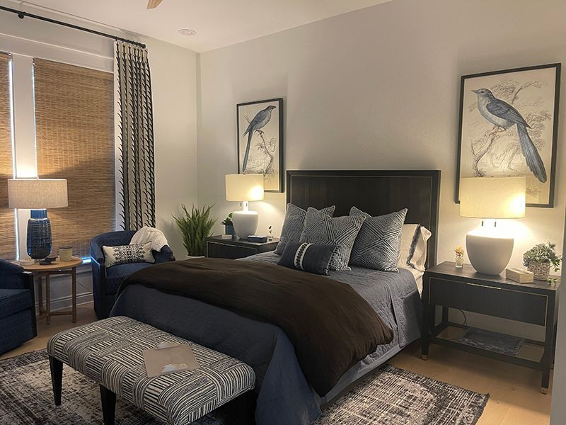 Cozy bedroom with a bed, patterned bench, bedside lamps, framed bird art, and a blue armchair by the window