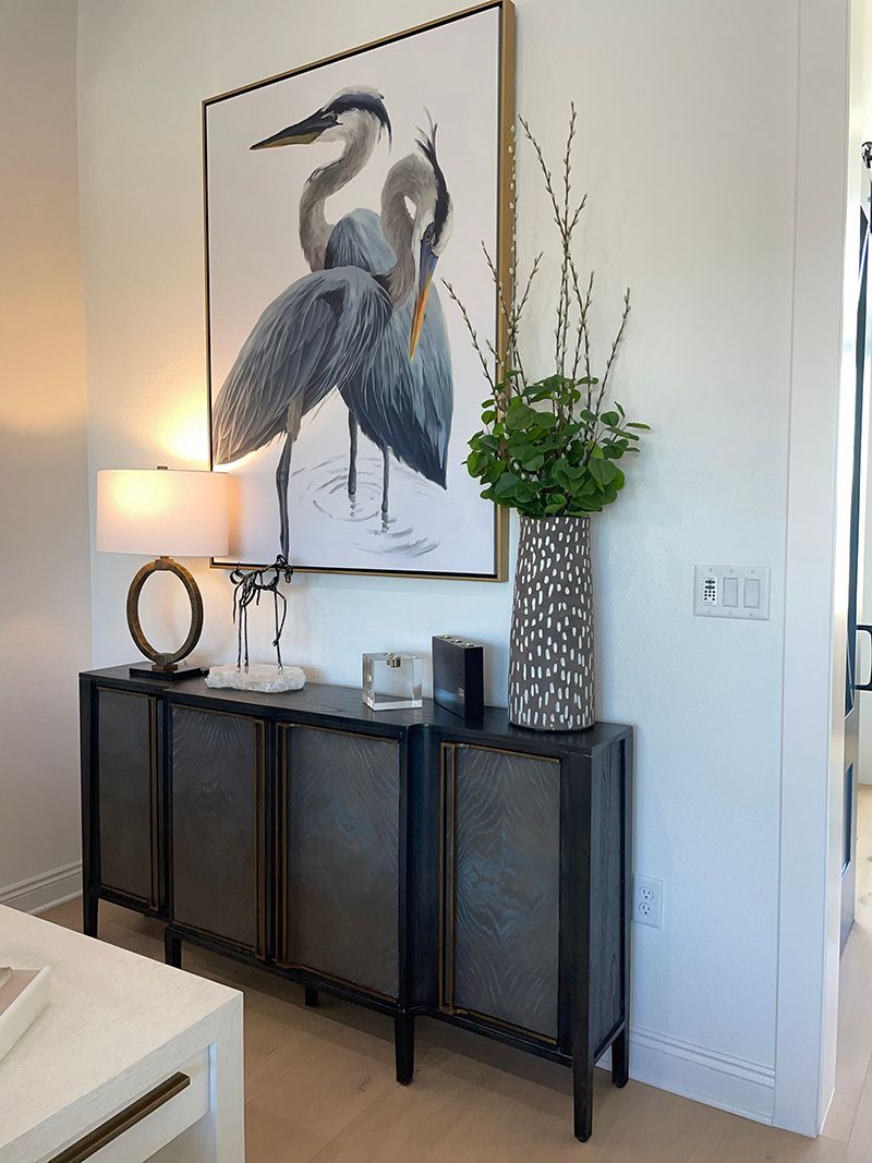 Modern console table with lamp, art print of herons, and tall vase of greenery in a bright room
