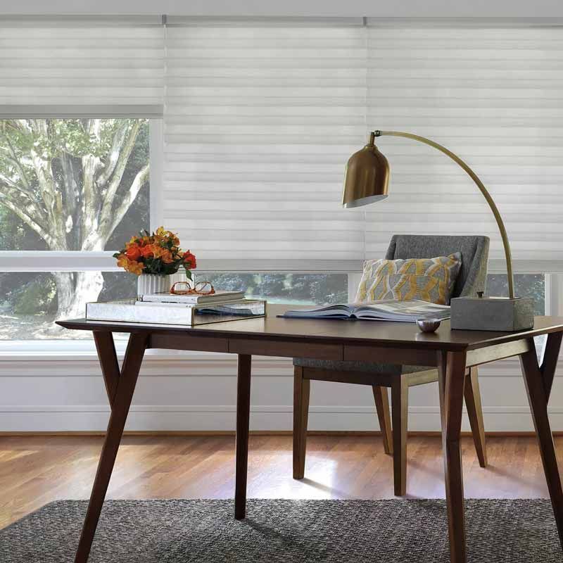 Modern home office with wooden desk, chair, brass lamp, flowers, and large window with blinds