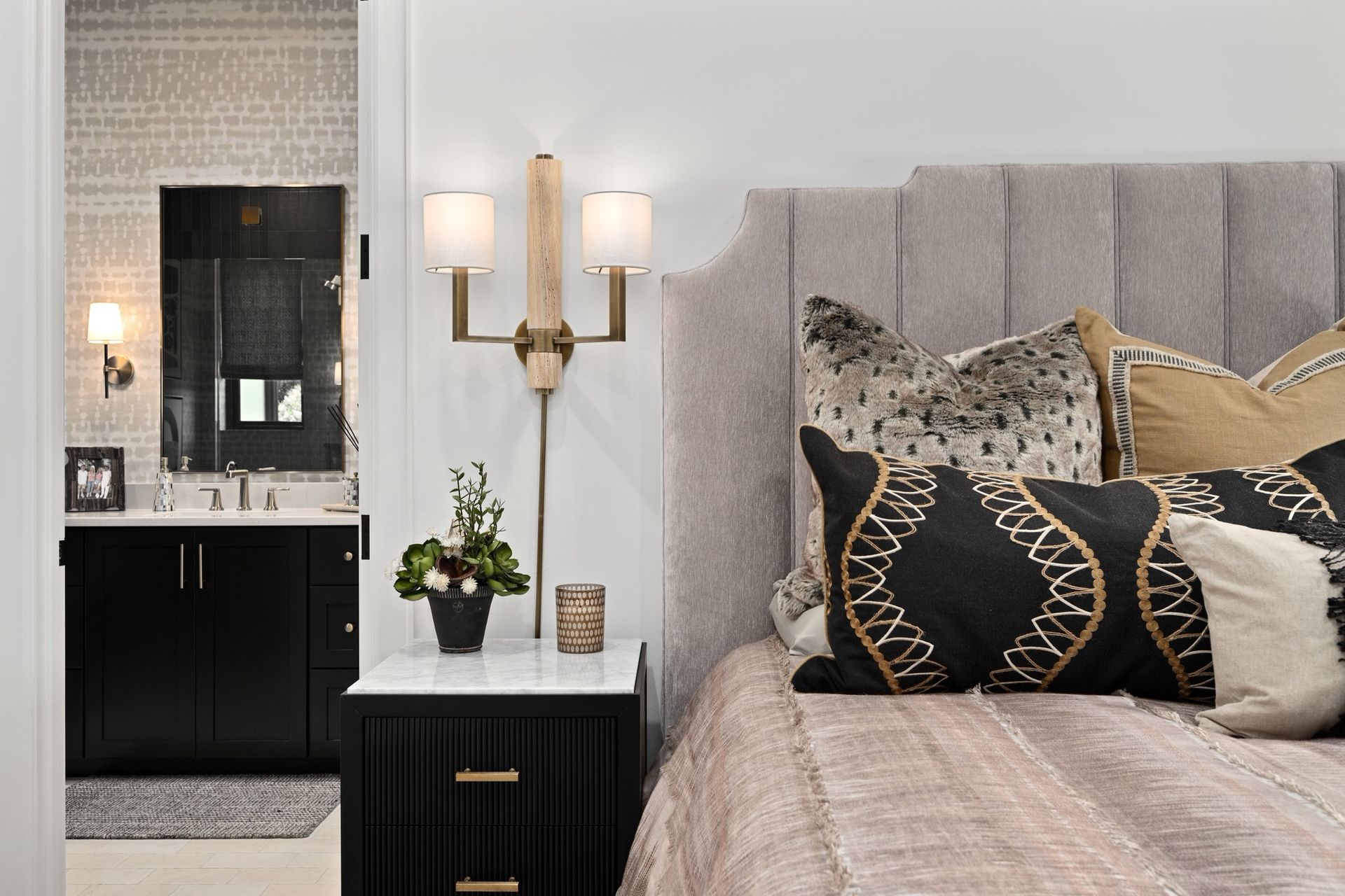 Elegant bedroom with tufted gray headboard, black nightstand, gold lamp, and mirrored vanity in the background