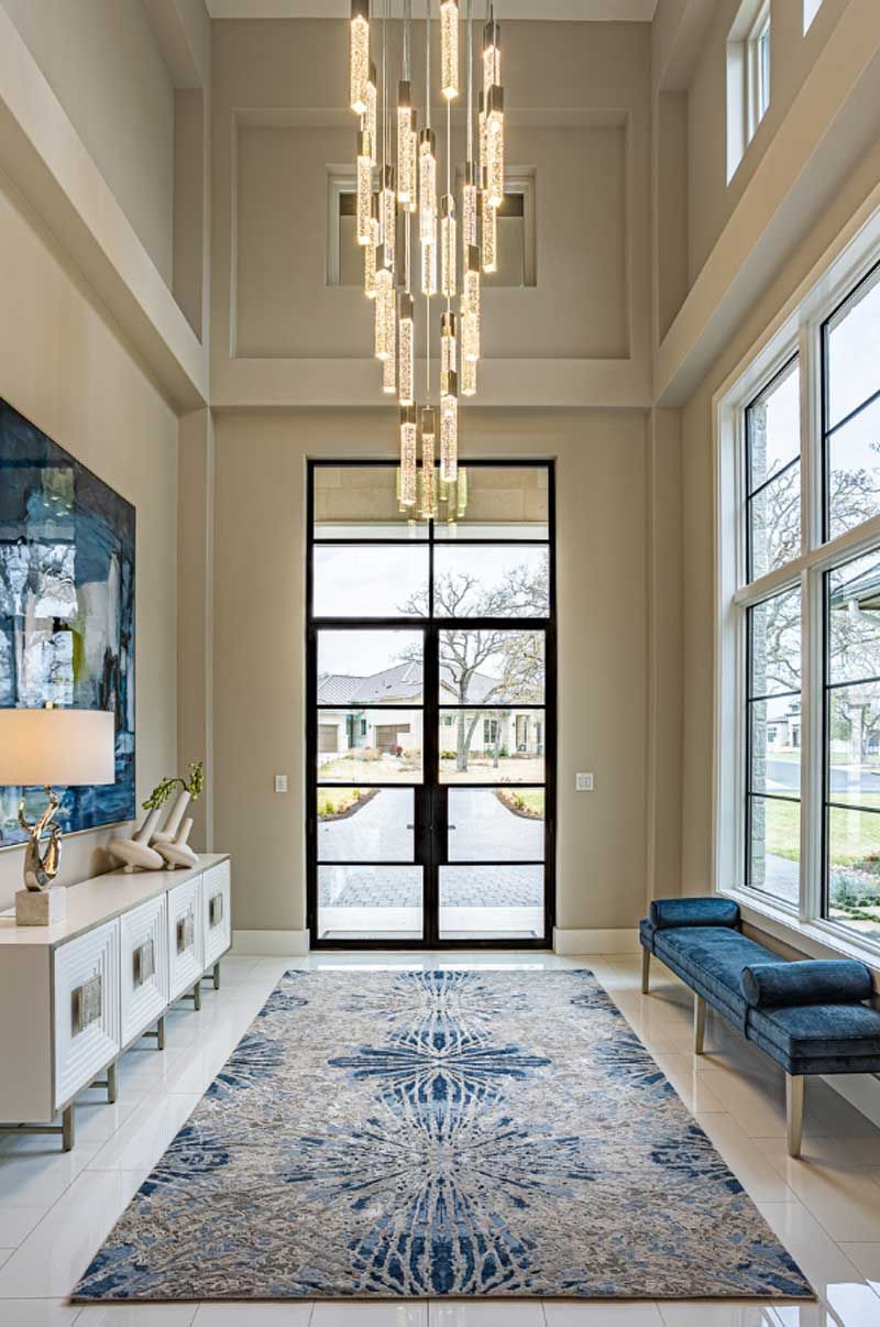 Bright modern foyer with tall windows, hanging chandelier, blue bench, and patterned rug