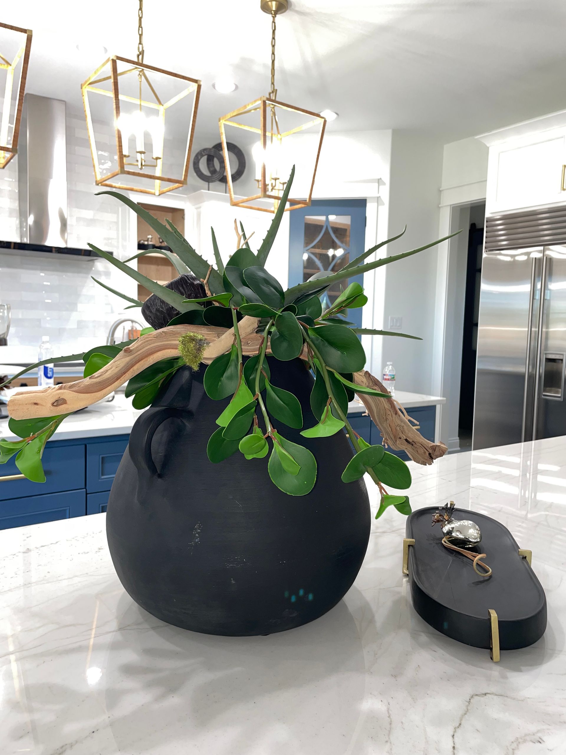Large black vase centerpiece with green plant on a kitchen island under gold pendant lights