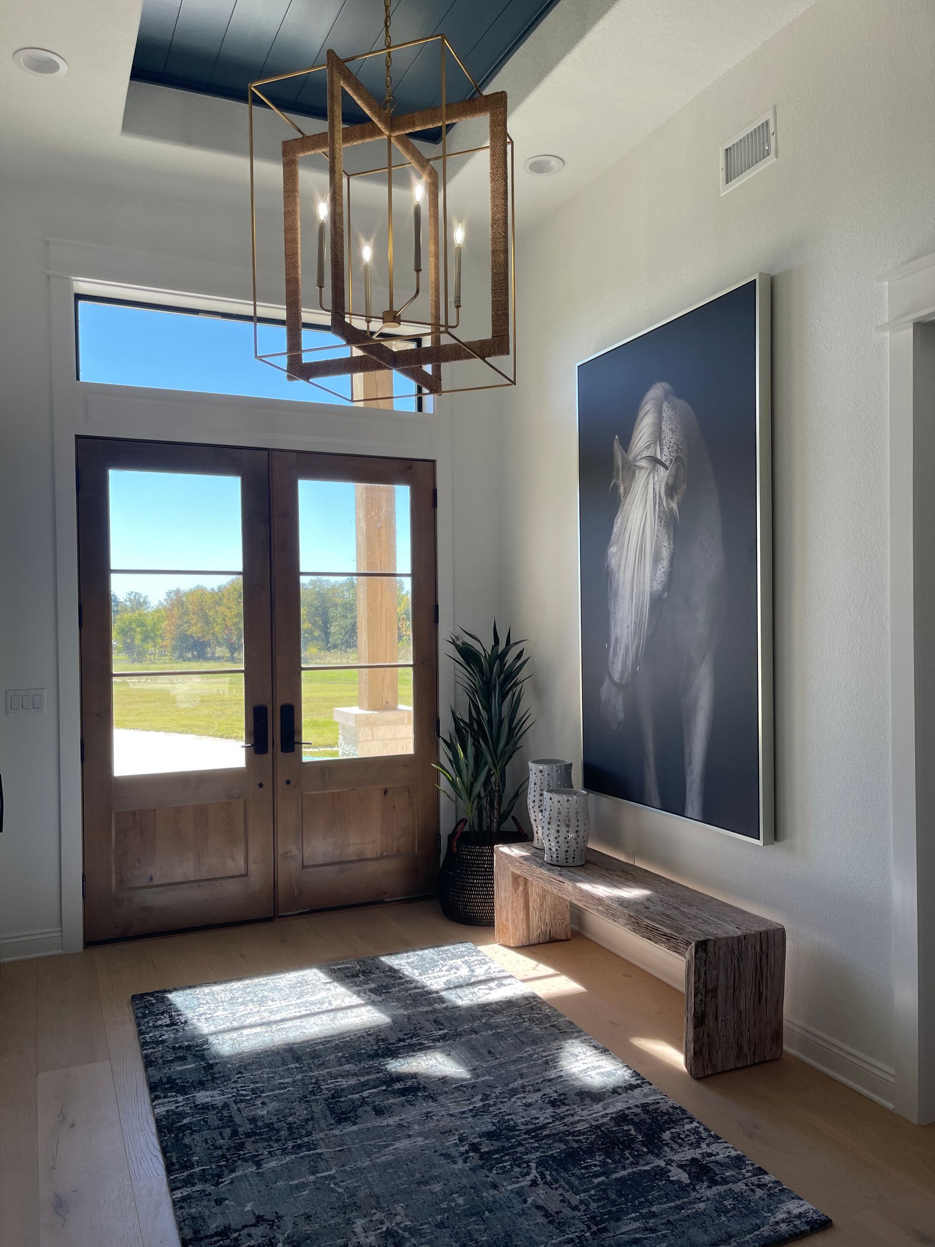 Bright entryway with wooden front doors, geometric pendant light, bench, large wall art, and patterned rug.