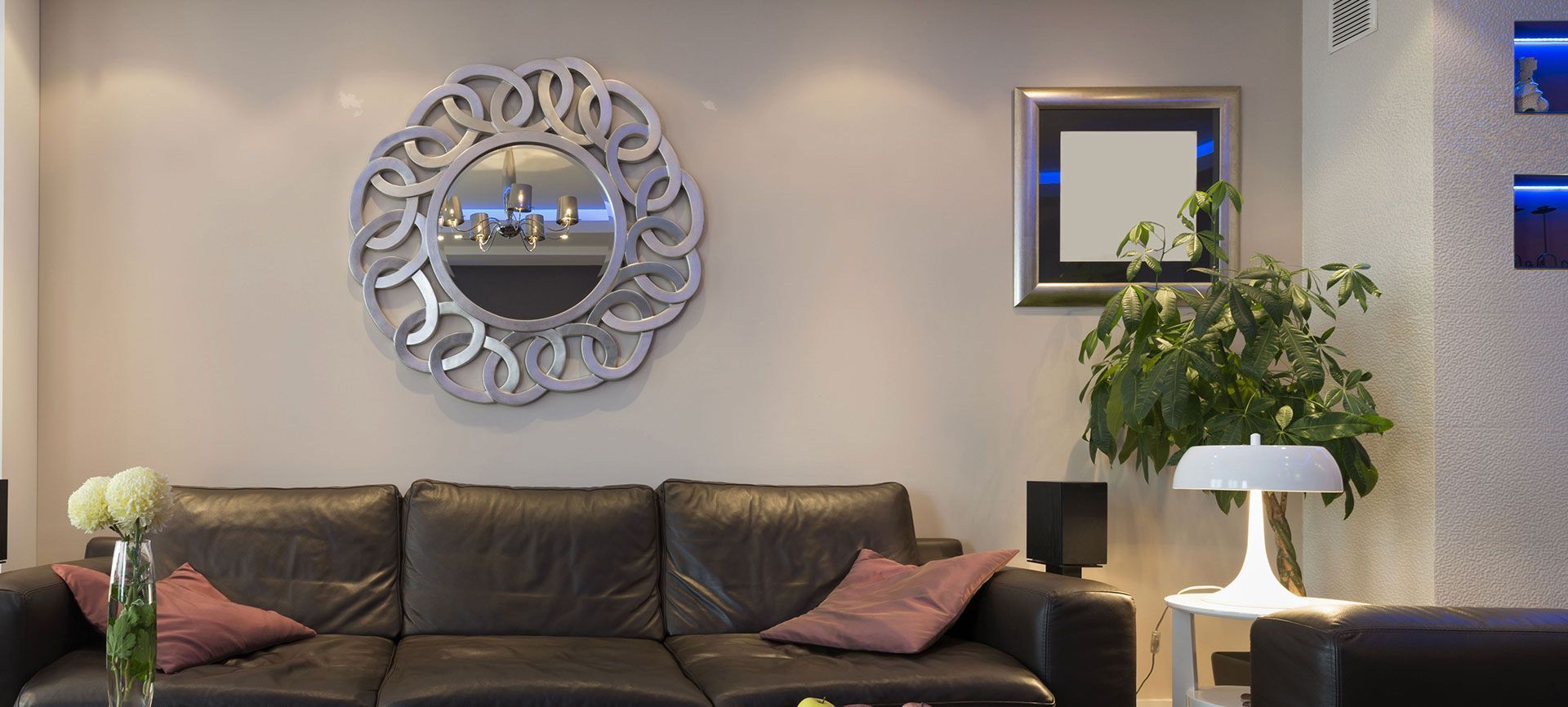 Modern living room with dark sofa, pink pillows, ornate wall mirror, framed art, and potted plant.