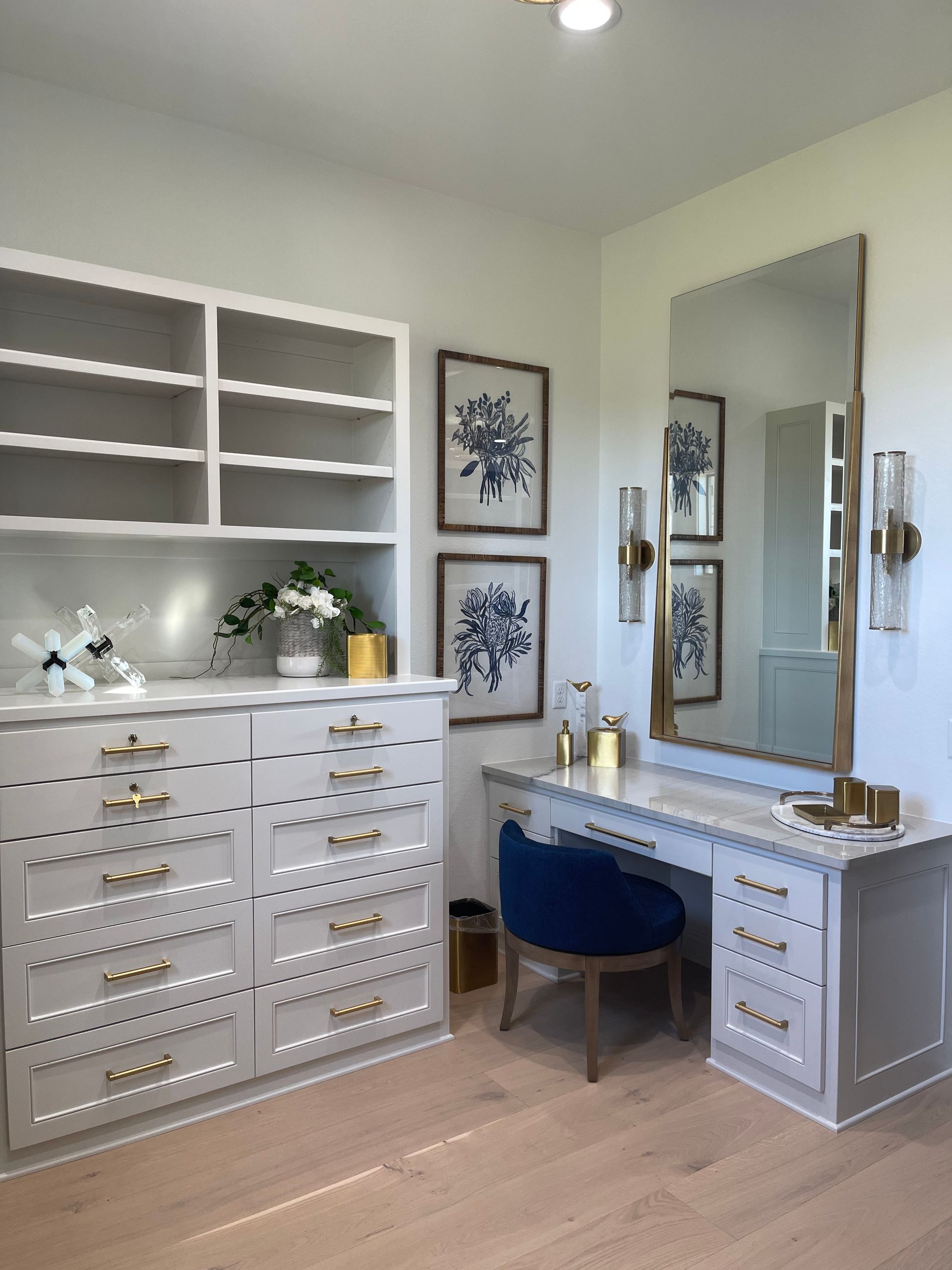 Elegant white vanity room with built-in shelves, drawers, mirror, and blue chair on a light wood floor.