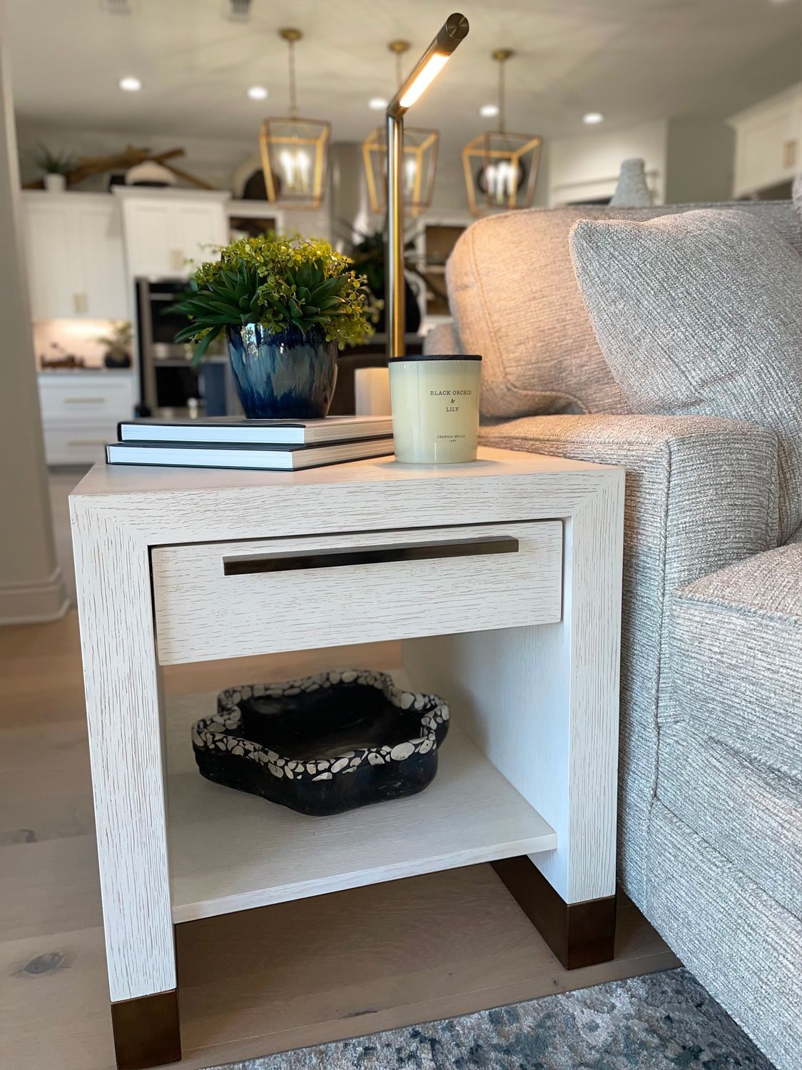 White woven side table beside a gray sofa, with books, a candle, a plant, and a black bowl shelf below.