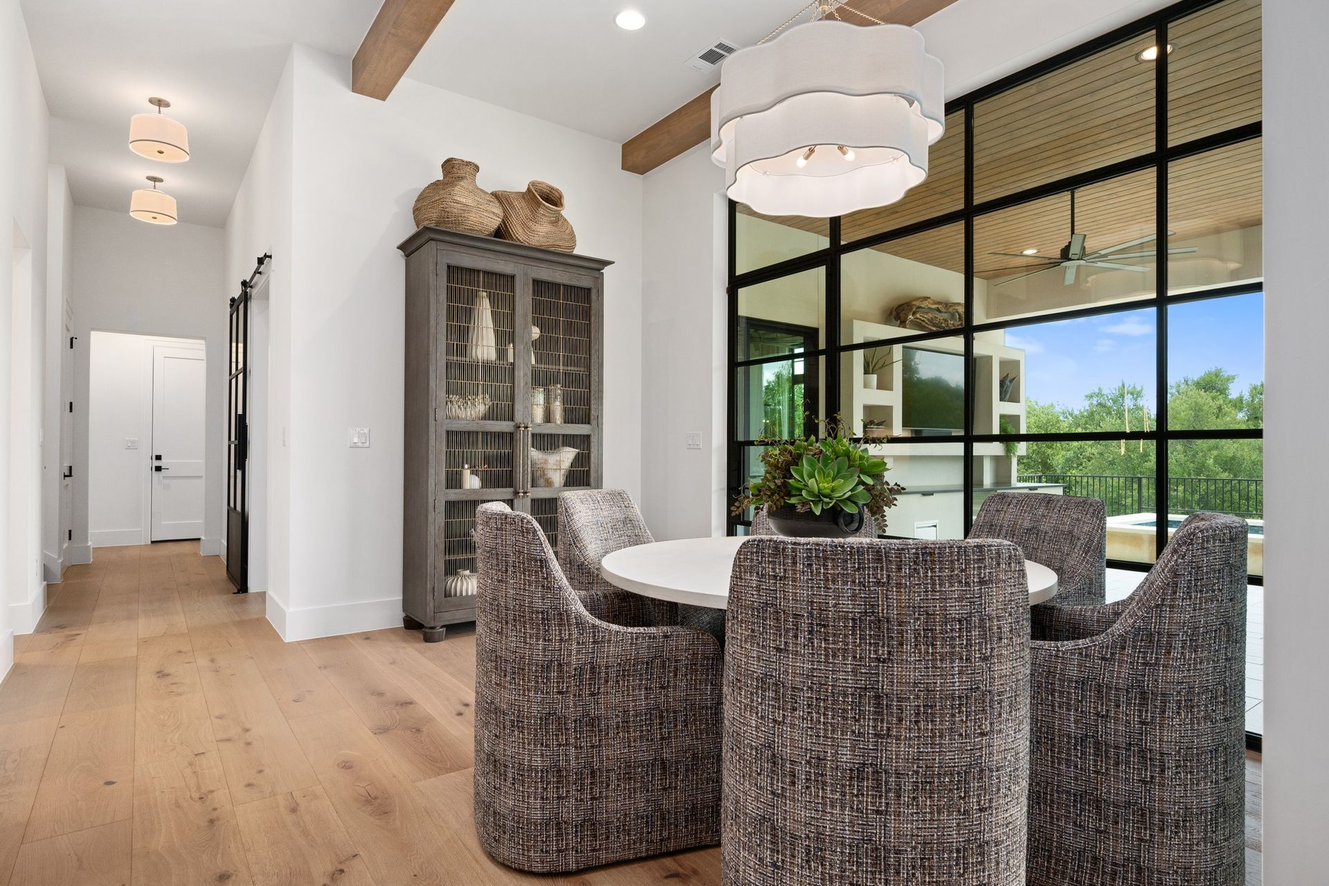 Bright dining room with woven chairs, round table, glass cabinet, and floor-to-ceiling windows overlooking greenery