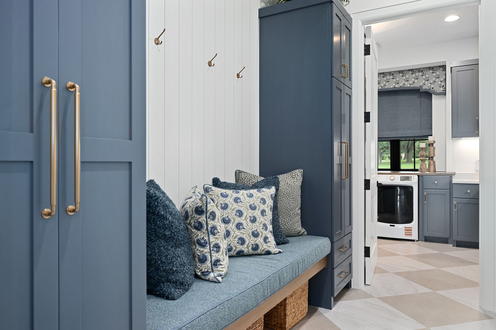 Blue mudroom bench with patterned pillows leading into a bright kitchen with gray cabinets and a window seat