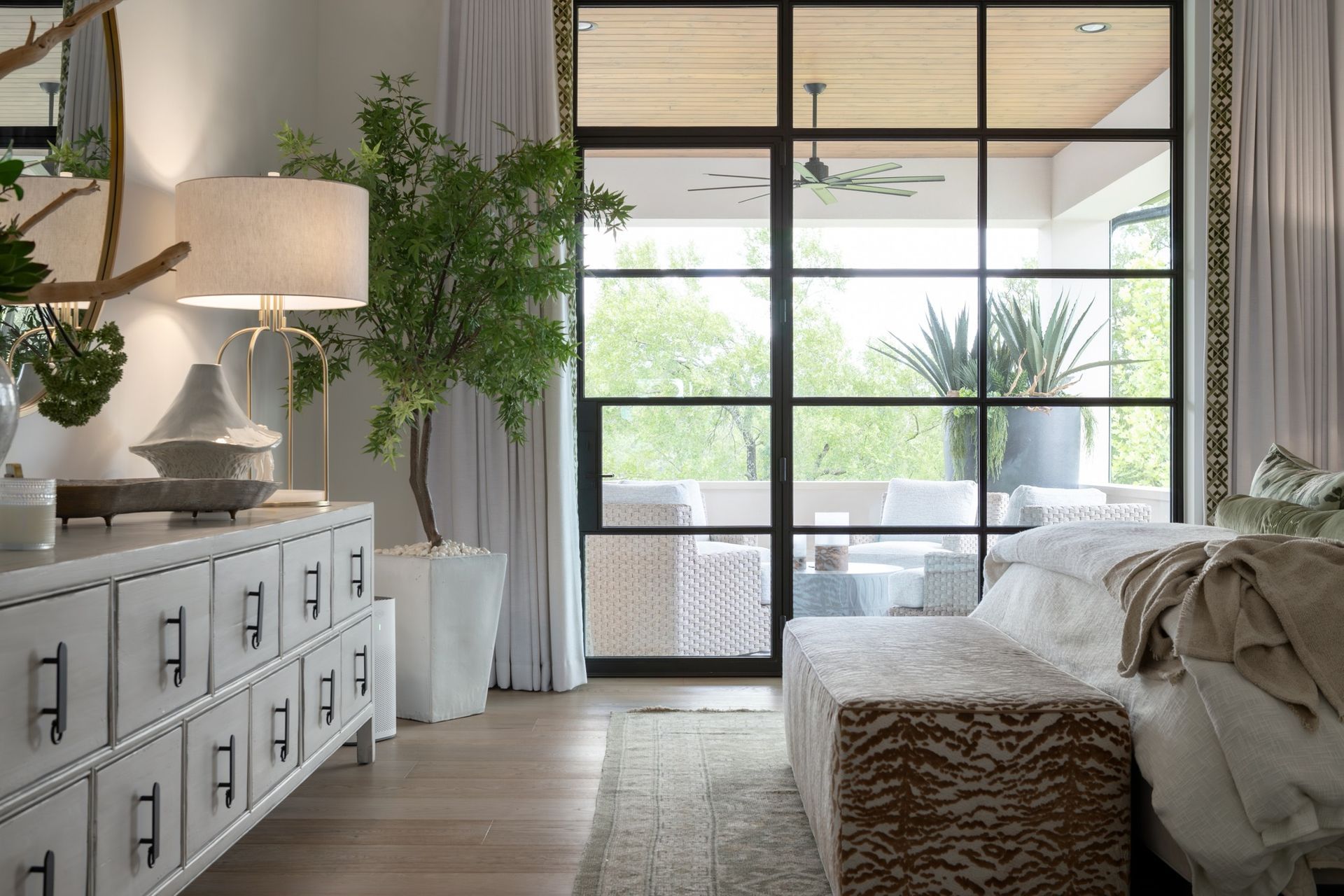 Bright bedroom with a bed, white dresser, hanging plants, and a glass wall opening to greenery
