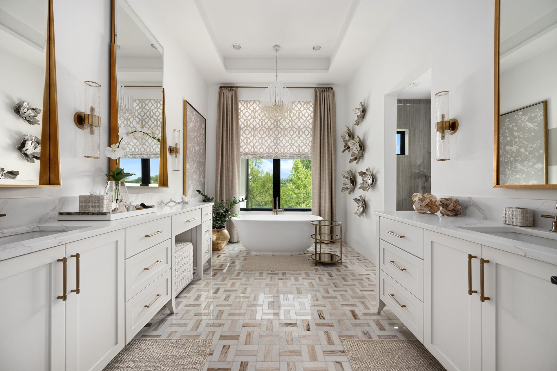 Bright, elegant bathroom with double vanities, gold accents, and a window seat overlooking greenery