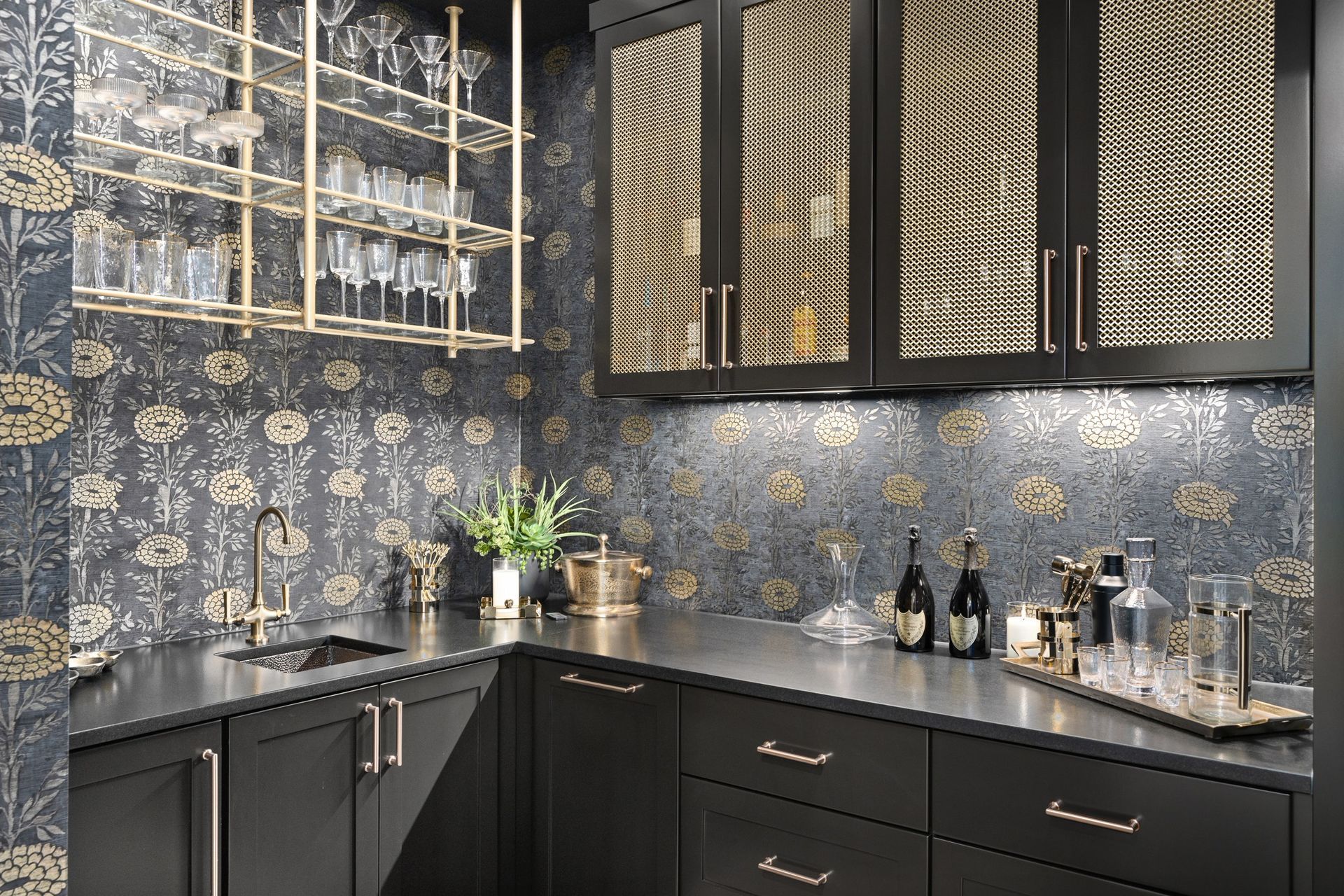 Stylish dark kitchen with floral wallpaper, black cabinets, gold accents, and bottles on the counter.