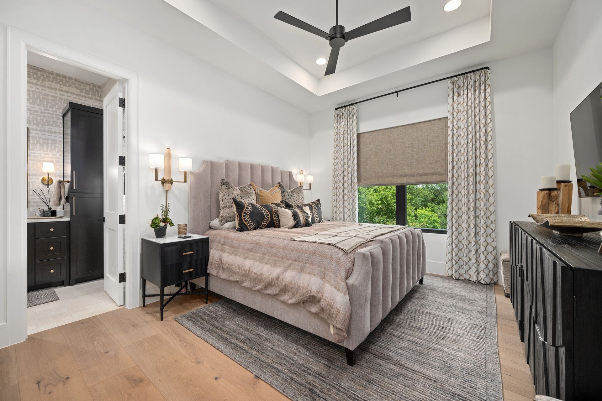 Modern bedroom with gray upholstered bed, black dresser, and large window with patterned curtains