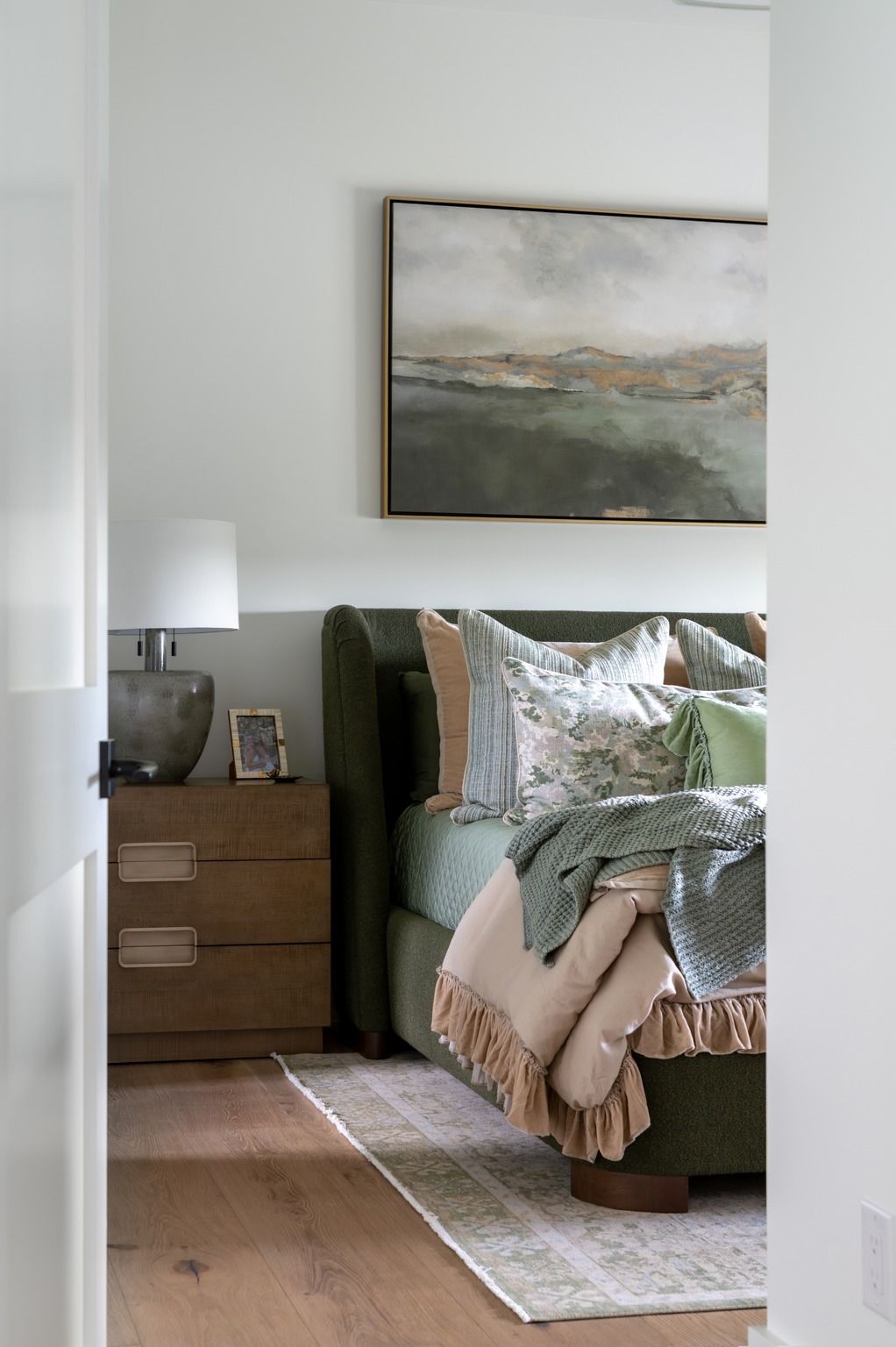 Cozy bedroom with a green bed, wooden nightstand, framed landscape art, and soft neutral bedding