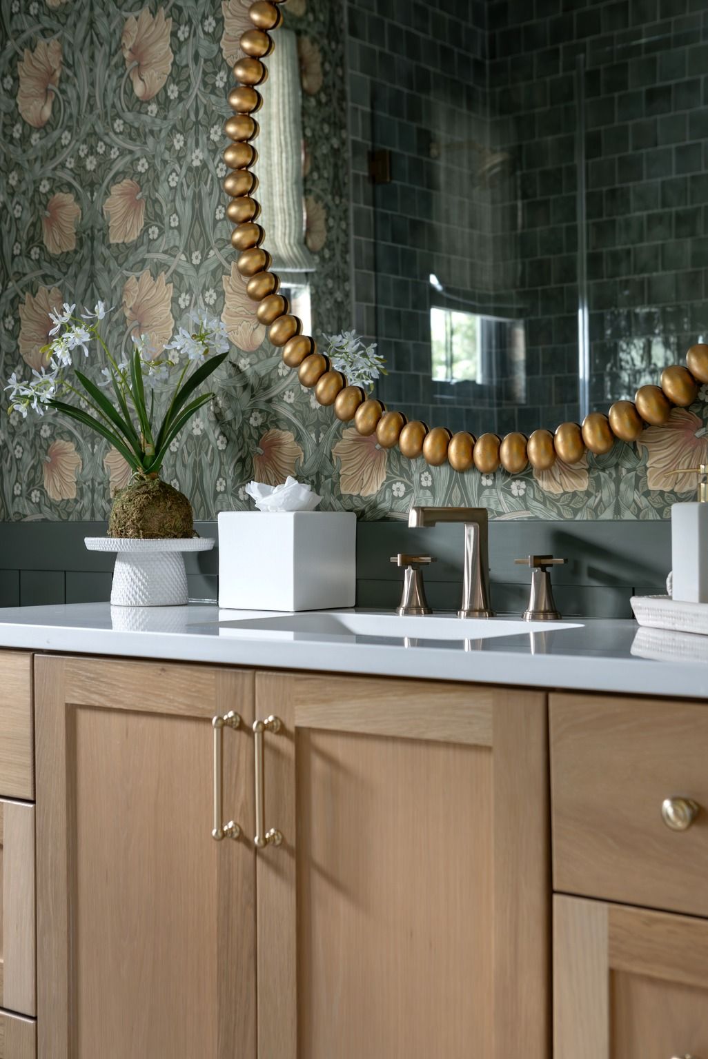 Bathroom vanity with wood cabinets, white countertop, round mirror, and small plants against green tile wall