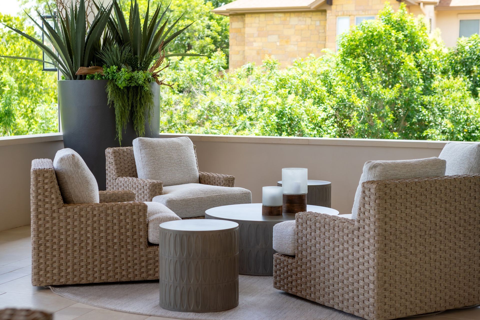 Wicker patio seating with beige cushions around a round table on a sunlit balcony, with potted plants nearby