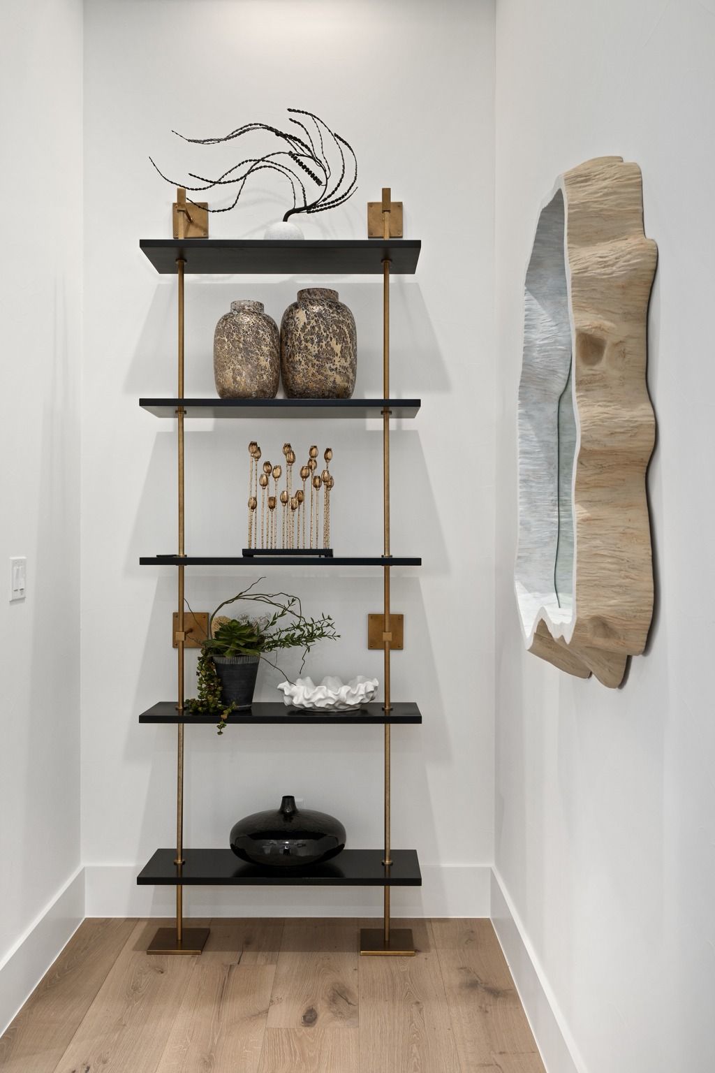 Tall black shelf with decor and a stone wall mirror in a minimalist hallway.