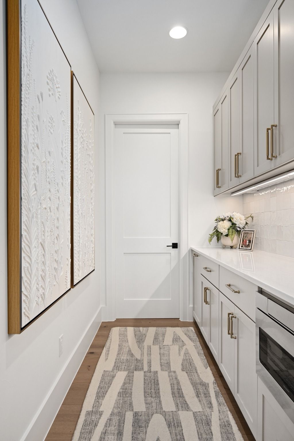 Bright narrow galley kitchen with white cabinets, patterned runner, and closed door at the end