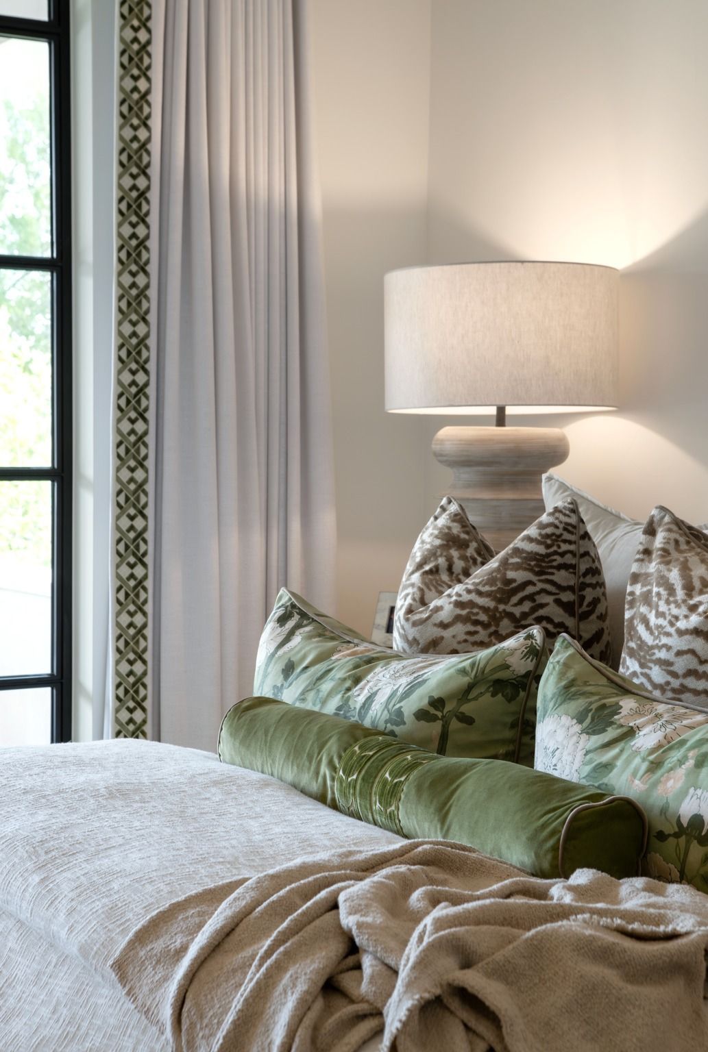 Cozy bed with green pillows, beige throw, and a lit lamp beside a window