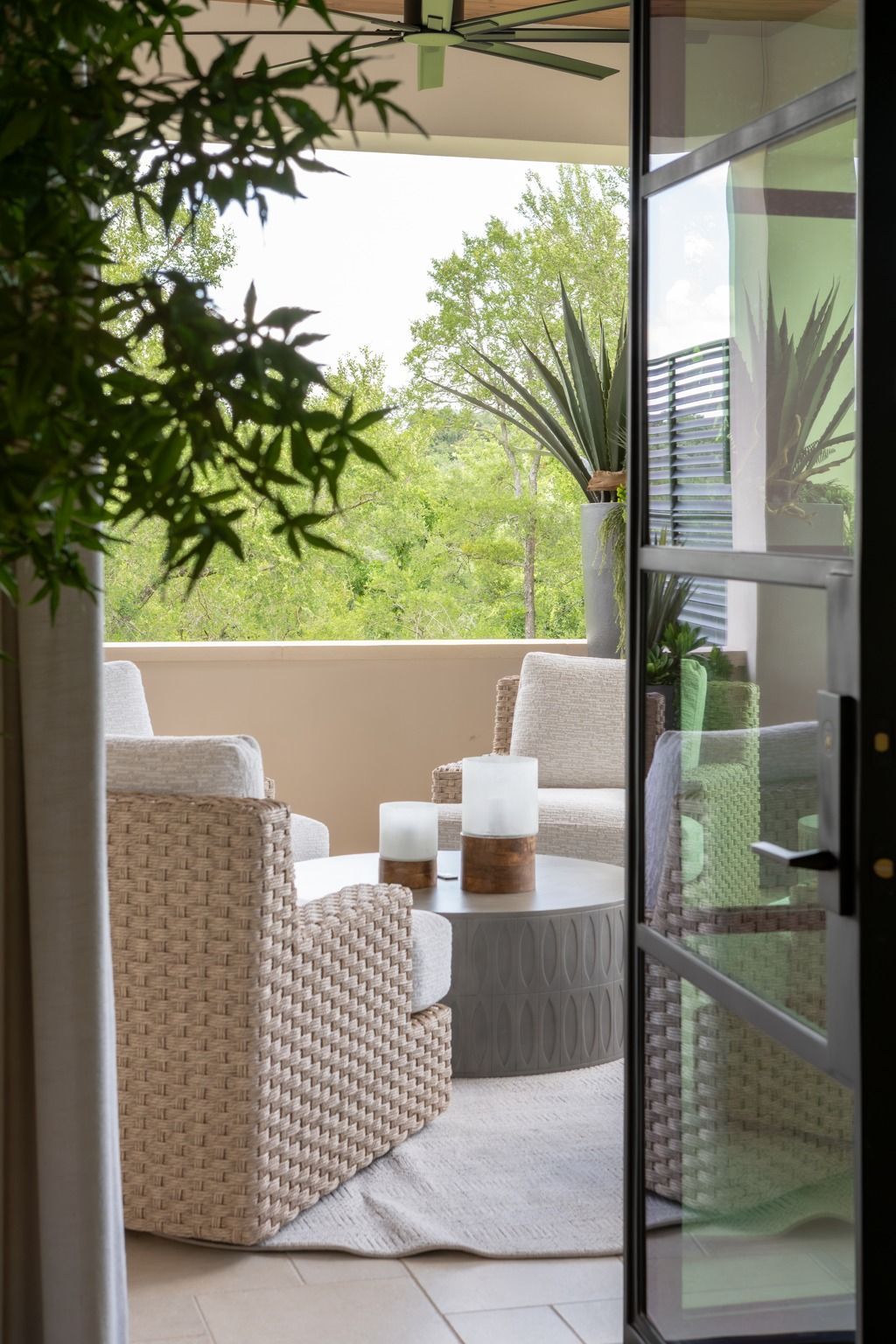 Open balcony with wicker chairs, round table, and lush greenery beyond glass doors