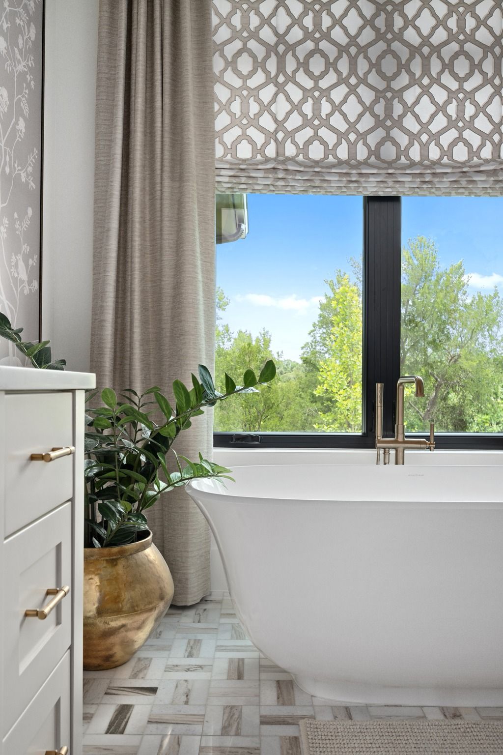 Bright bathroom with freestanding tub, patterned shades, and large window overlooking green trees