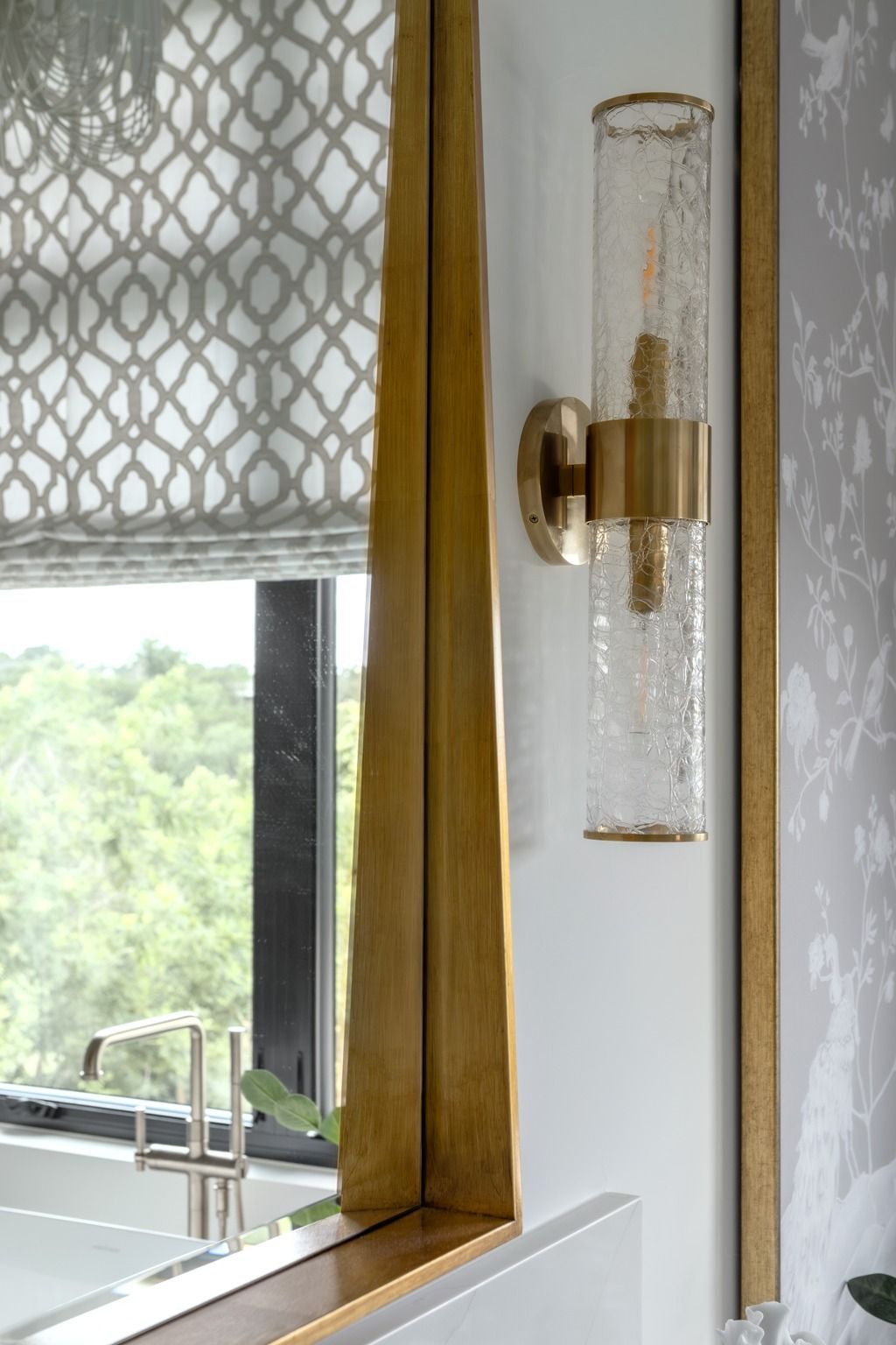 Gold wall sconce beside a window with patterned blinds and a view of greenery