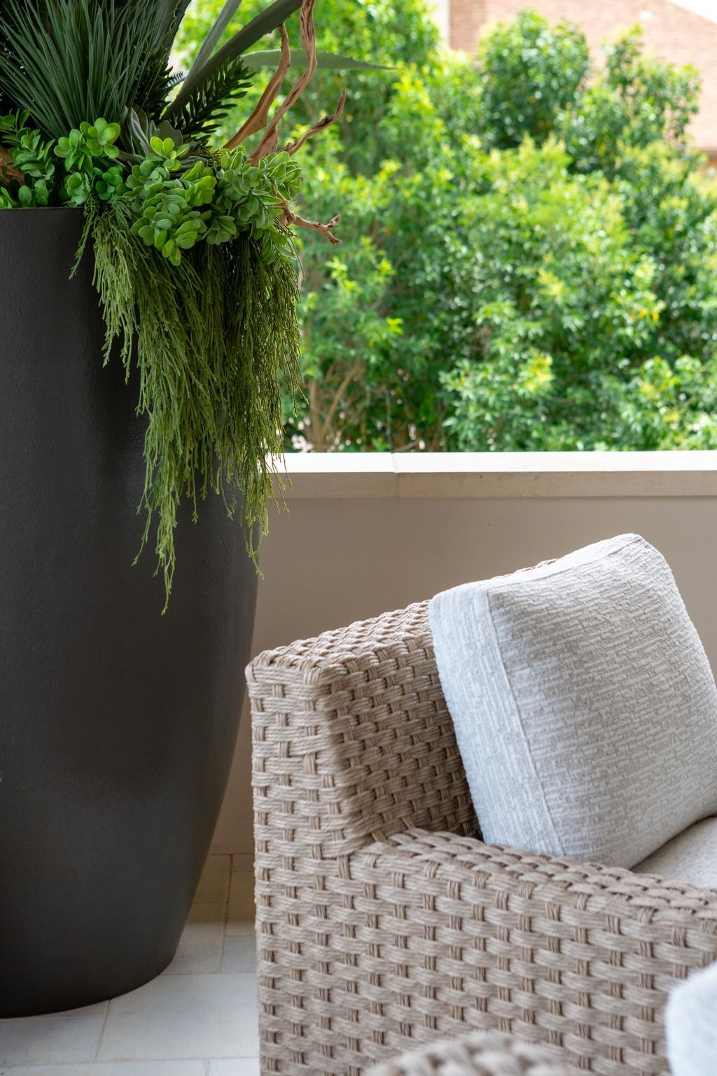 Woven chair with white cushion beside a tall black planter of cascading green ferns on a balcony.
