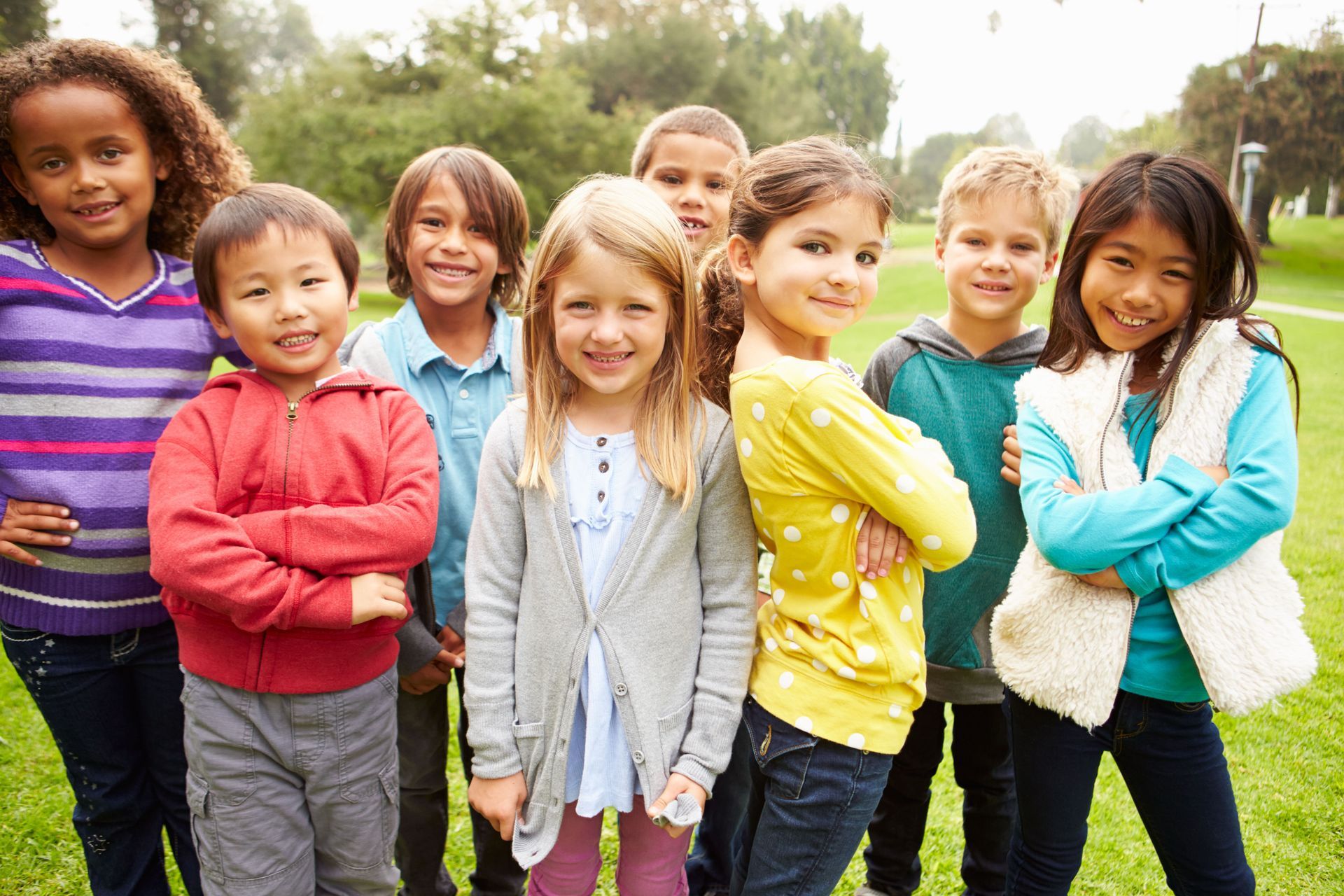 Group of diverse children smiling outdoors, some with arms crossed.