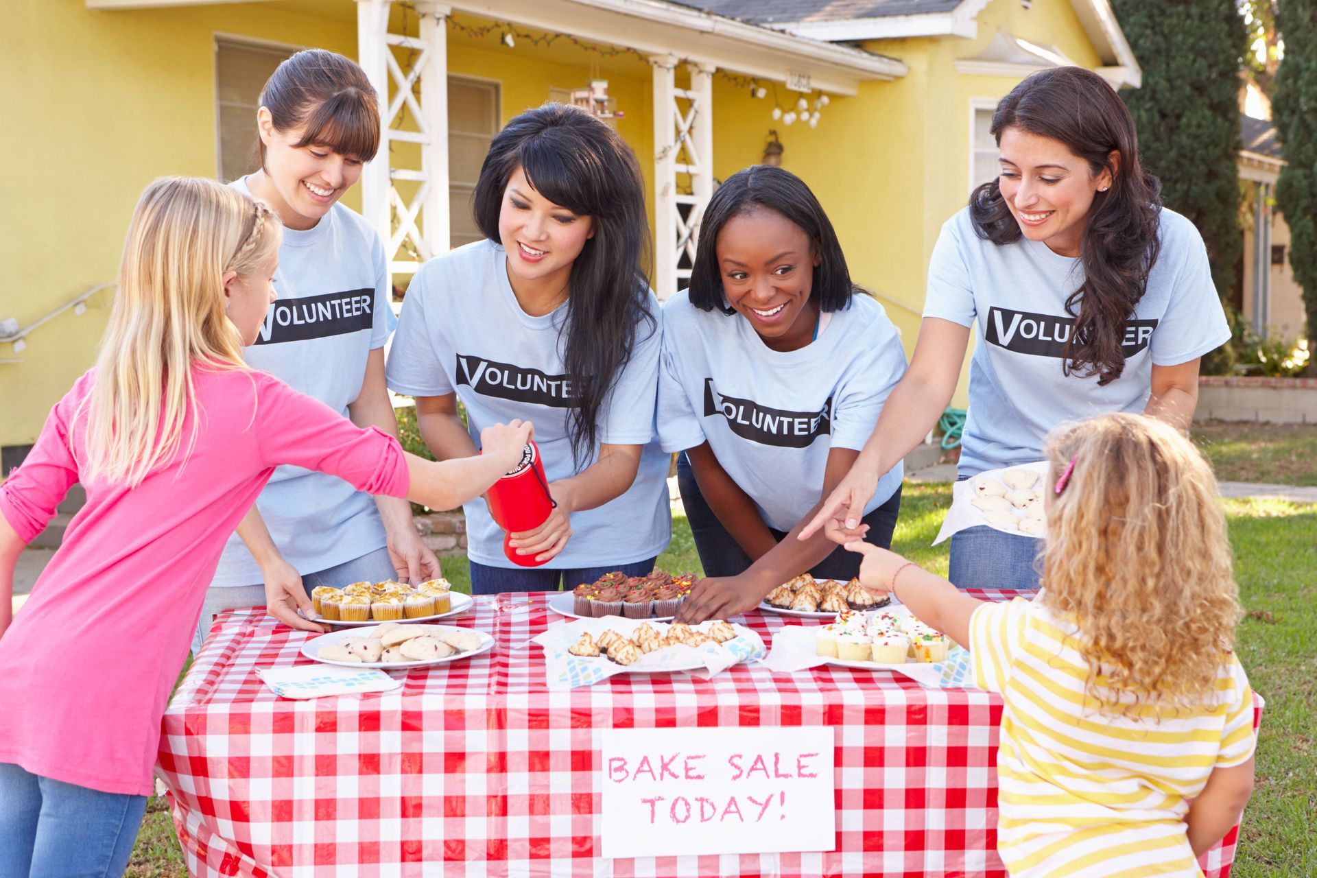 People at a bake sale table; volunteers in blue shirts help children select baked goods.