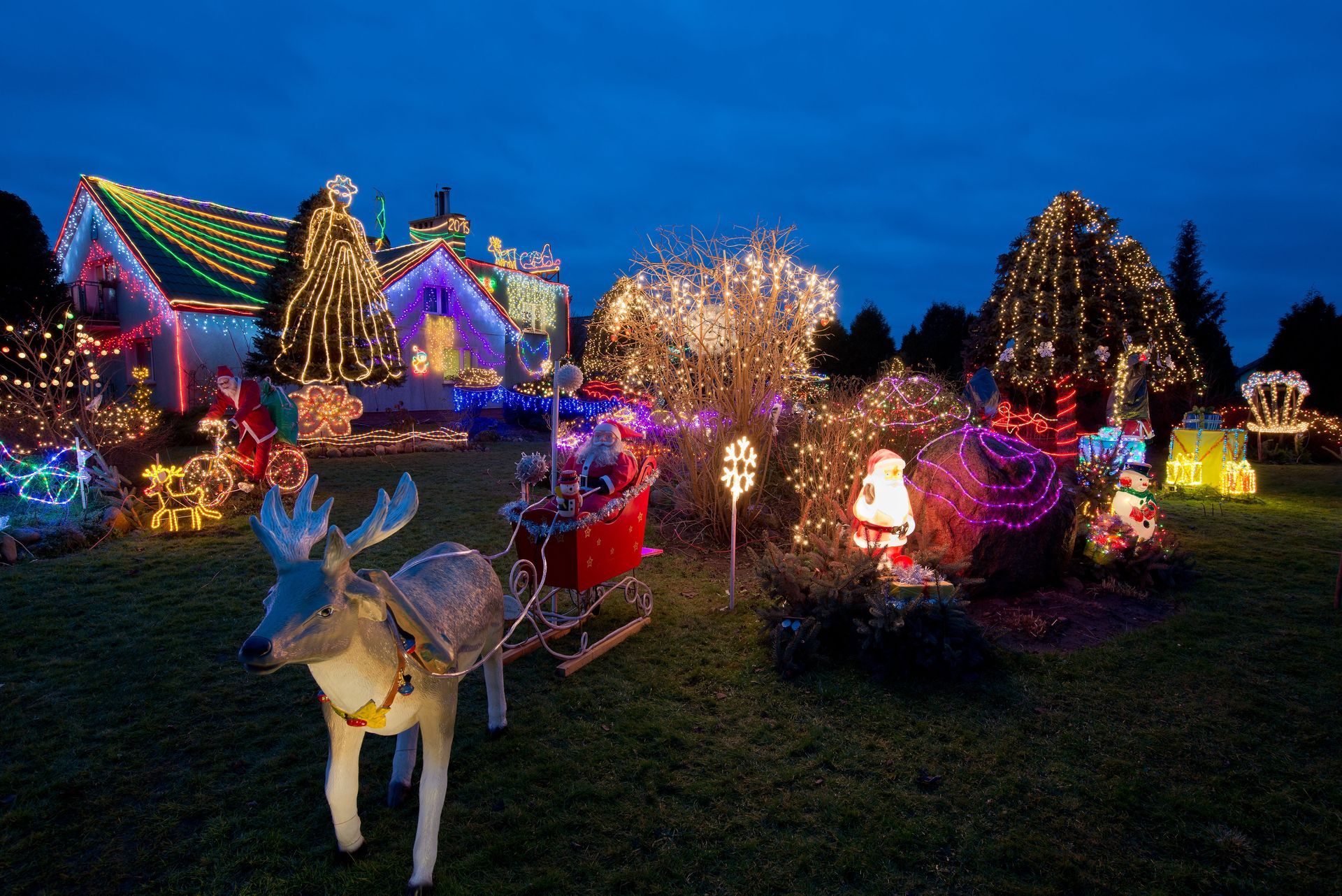 A house decorated with Christmas lights, including a reindeer, Santa, and a sleigh, at dusk.