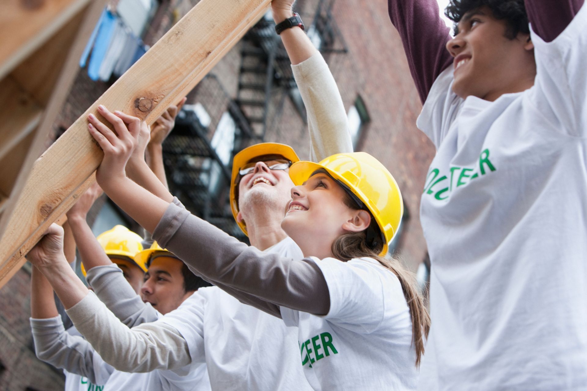 Volunteers lifting wood beam, smiling, wearing hard hats and