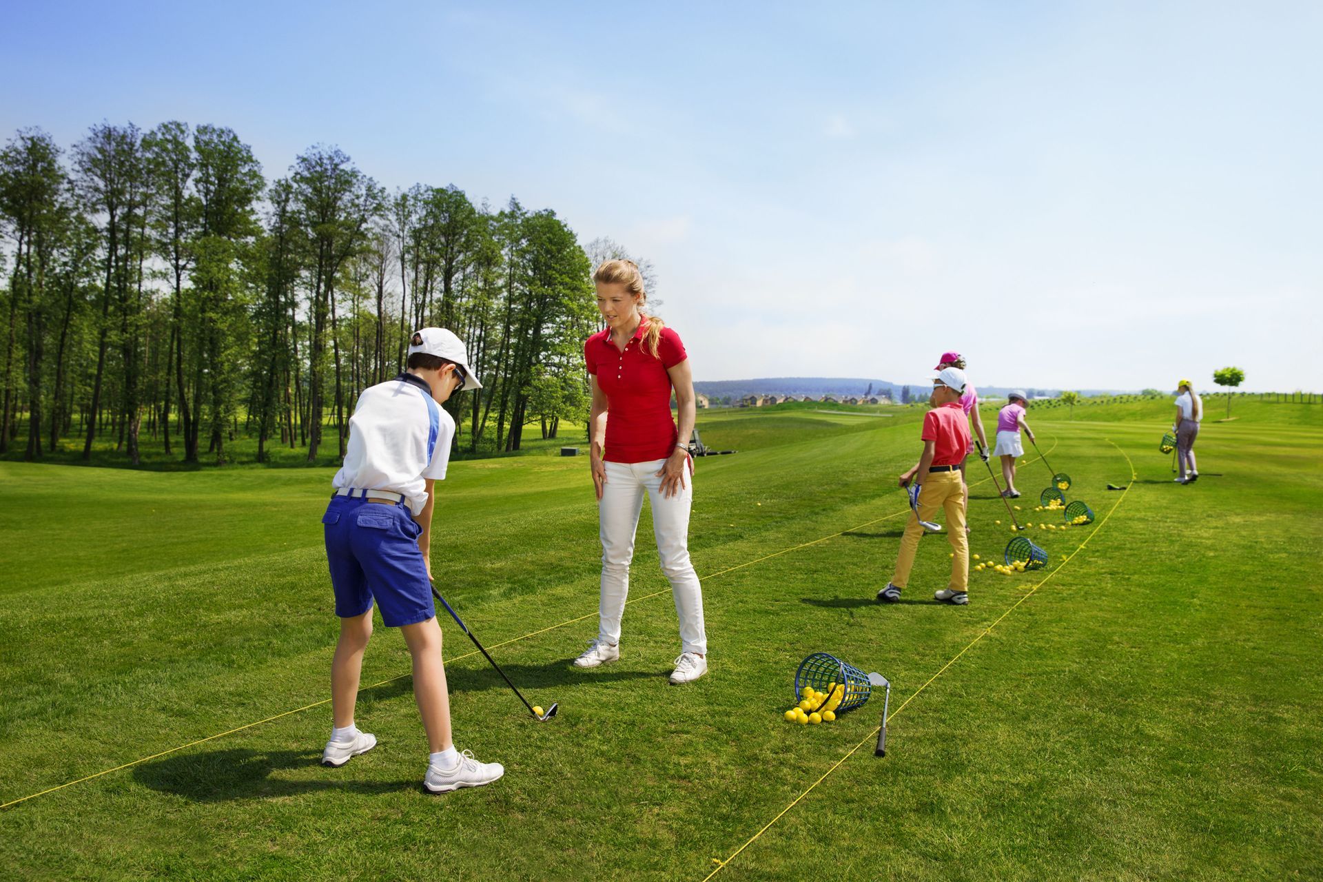 Boy taking a golf swing, being coached by a woman on a green, other golfers in background.
