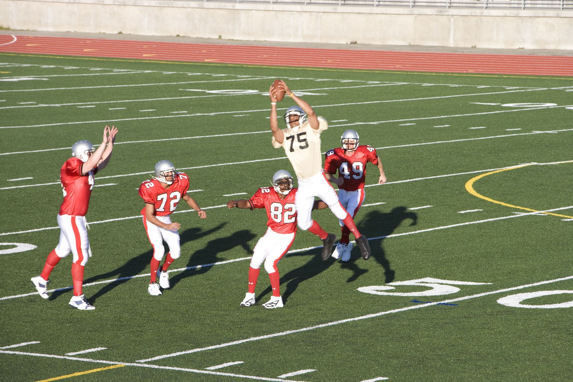 Football player in gold jersey jumps for a catch, surrounded by red-clad opponents on a green field.