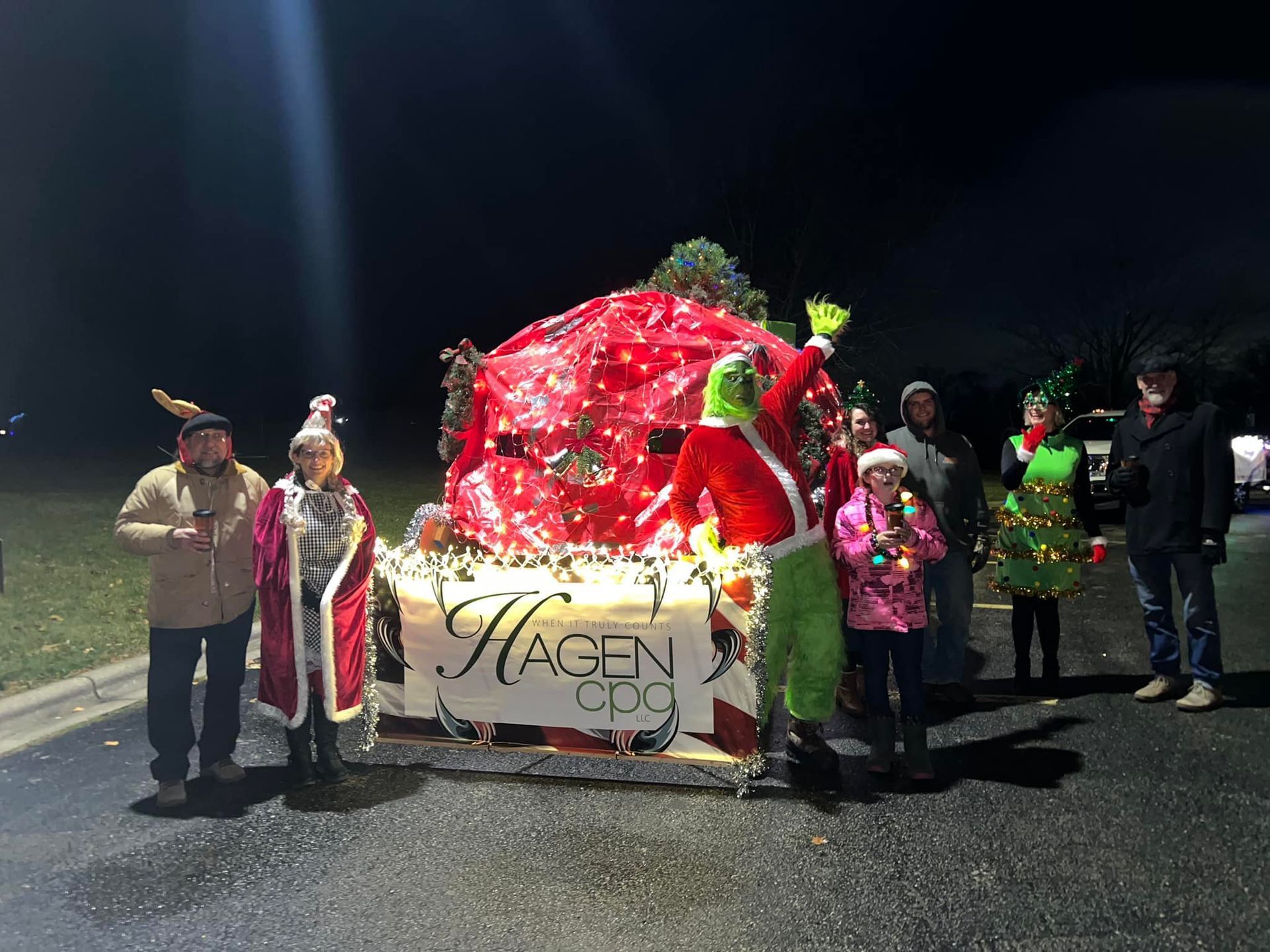 Parade float with Grinch and costumed people at night; red and white lights and a sign