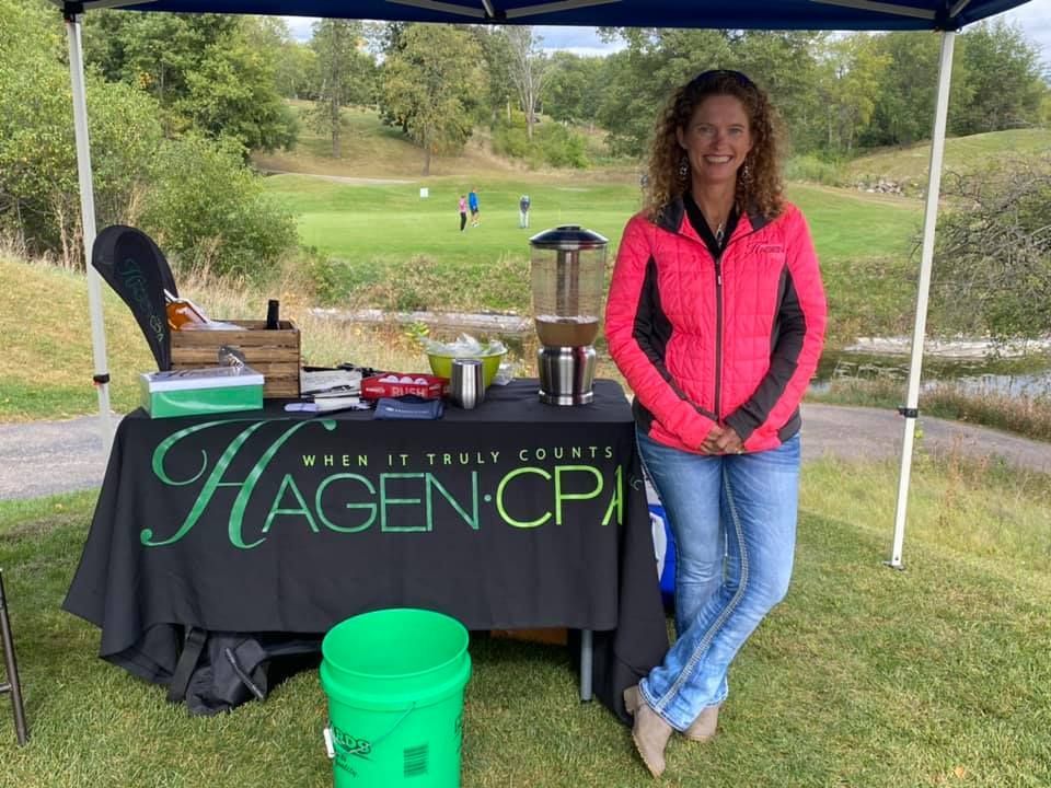 Woman in pink jacket at a Hagen CPA table on a golf course, smiling.