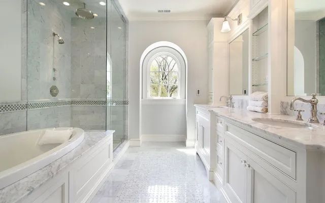 Bright white luxury bathroom with a soaking tub, glass shower, double vanity, and arched window.